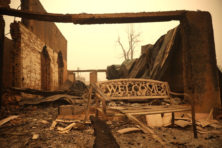 A welcome bench sits in front of the rubble of a building that was destroyed by the Dixie Fire. The sky is a yellow haze.