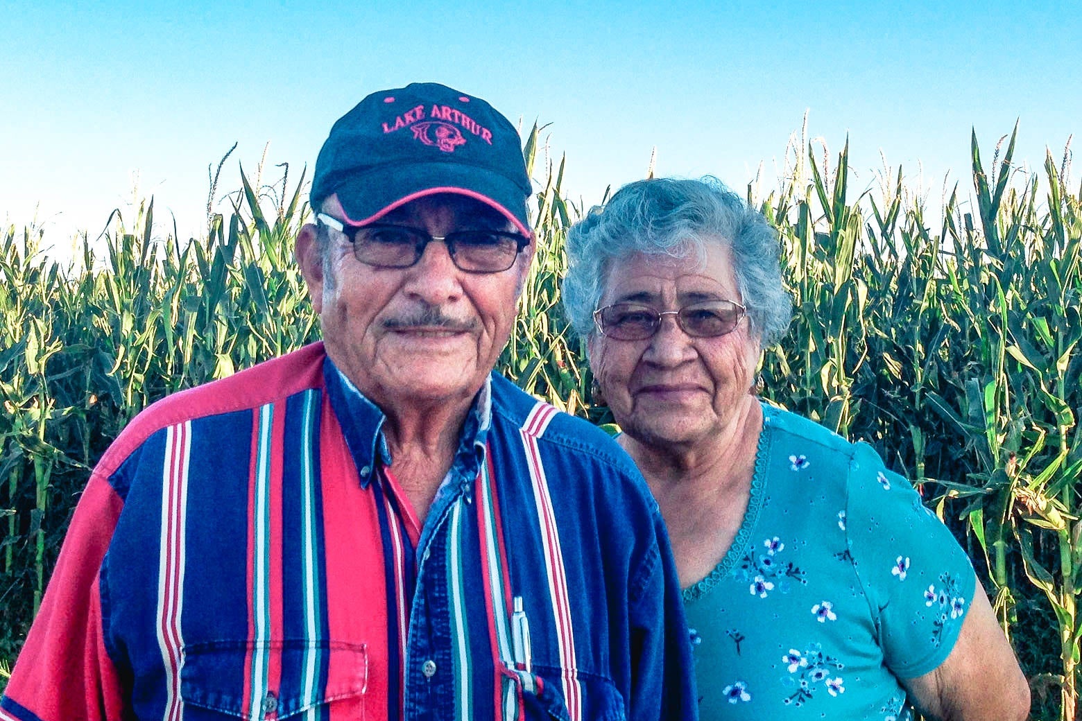 An older man and older woman stand in front of a corn field.