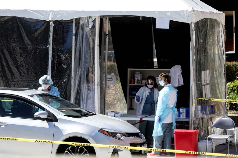 A doctor wearing a mask stands at a car parked by a plastic tent. Inside the tent, other doctors in masks stand in front of a shelf.