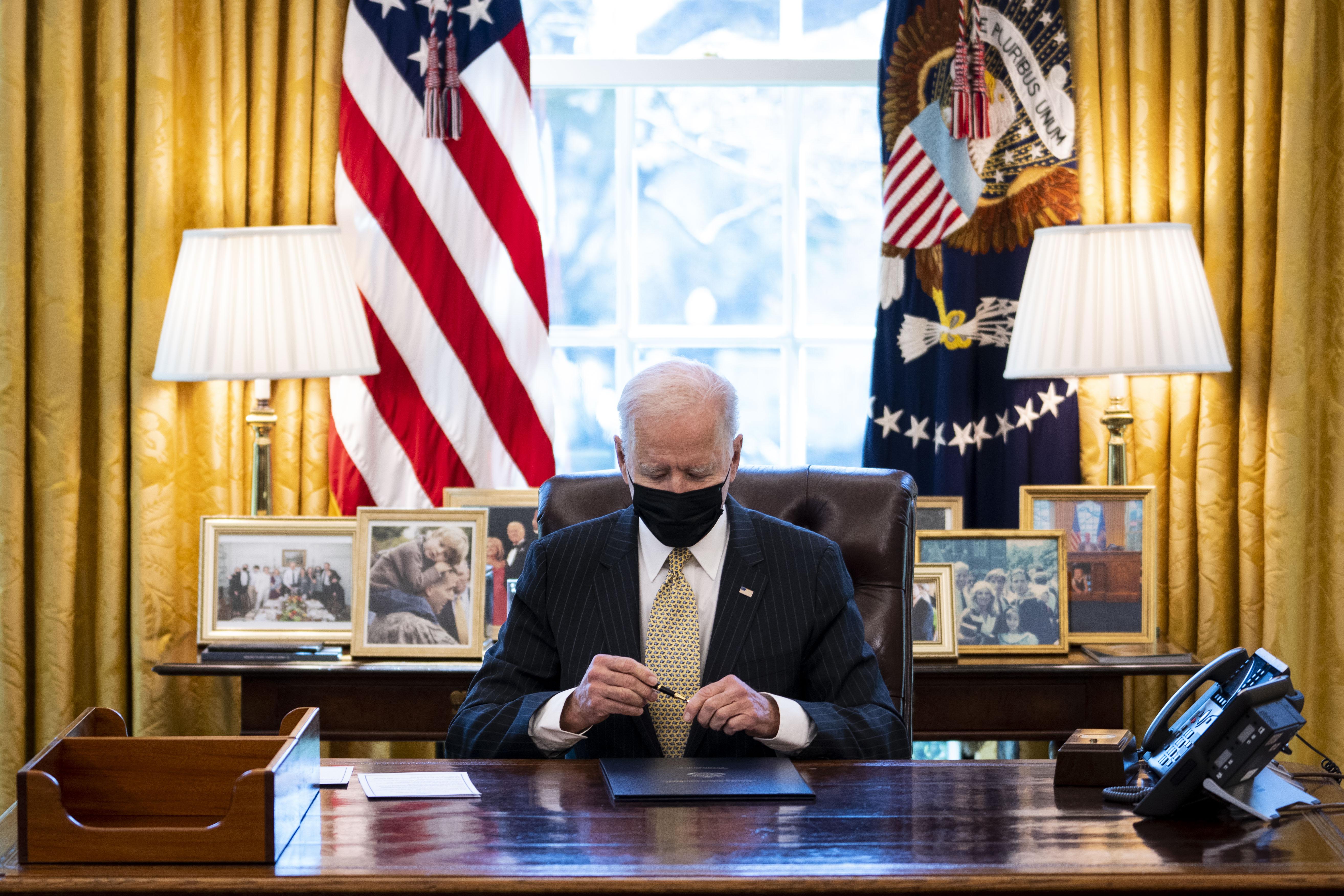 Biden caps a pen while seated at the Resolute Desk wearing a black mask