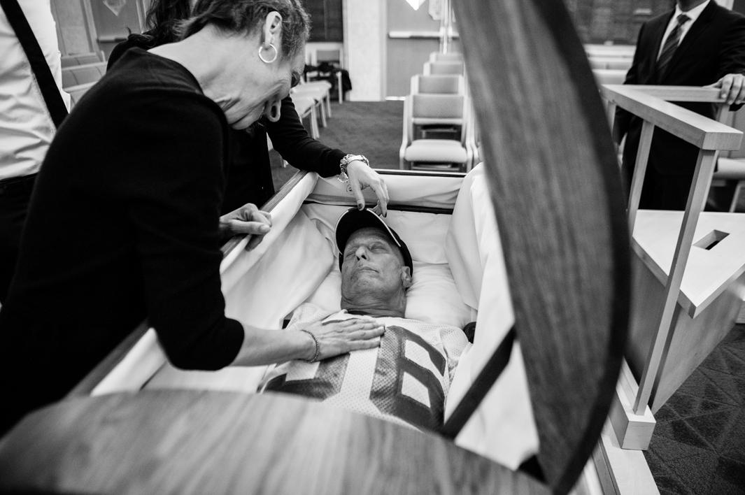 Howard Borowick left instructions for his funeral because he was a planner, and liked to be in charge, even in death. He requested to be buried in his favorite Giant’s football jersey, a pair of jeans, and his HB baseball cap. Laurel Borowick, his wife, smiles at him as they spend their last moment together before the casket is closed and the funeral begins. Mt. Kisco, New York. December 2013.