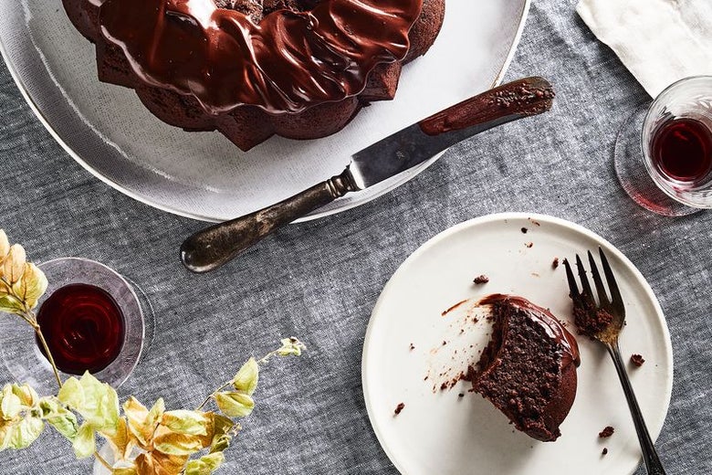 A slice of chocolate quinoa cake drenched in ganache sits on a plate, next to the whole cake and some Passover wine.