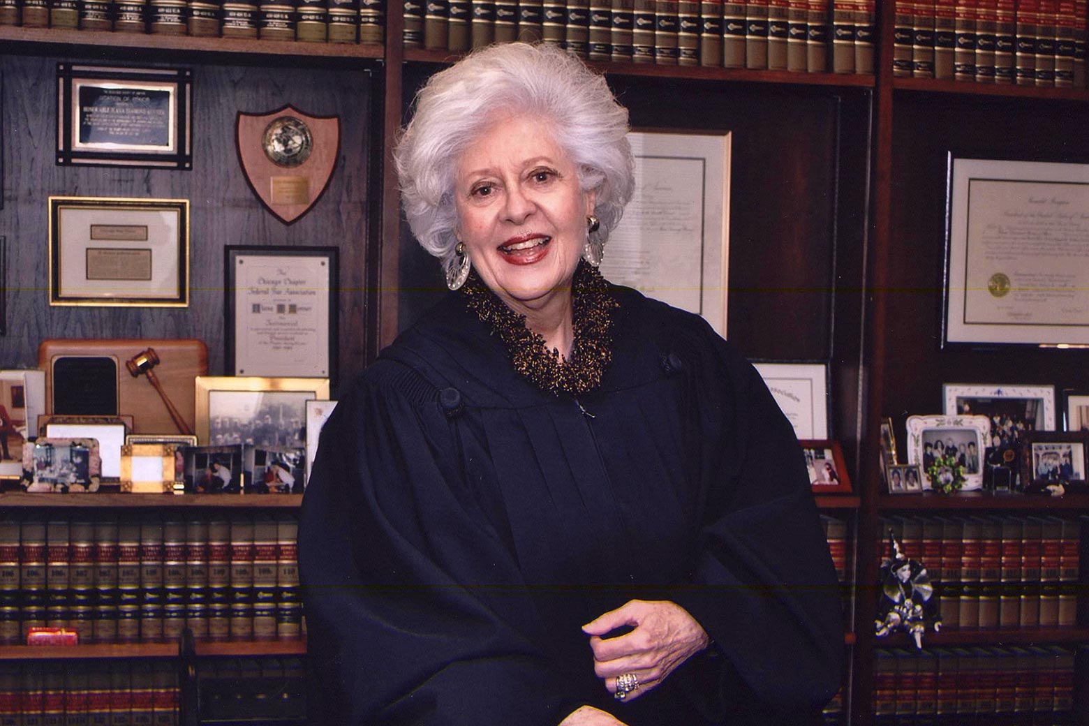 Judge Rovner stands in front of a bookcase with legal books and a gavel and wears her judicial robe, large earrings, and a smile.