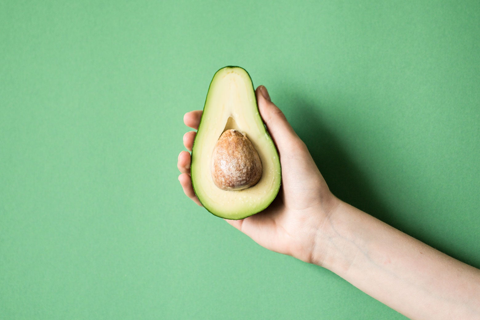 A hand holds up a perfectly ripe sliced avocado against an avocado-green background.