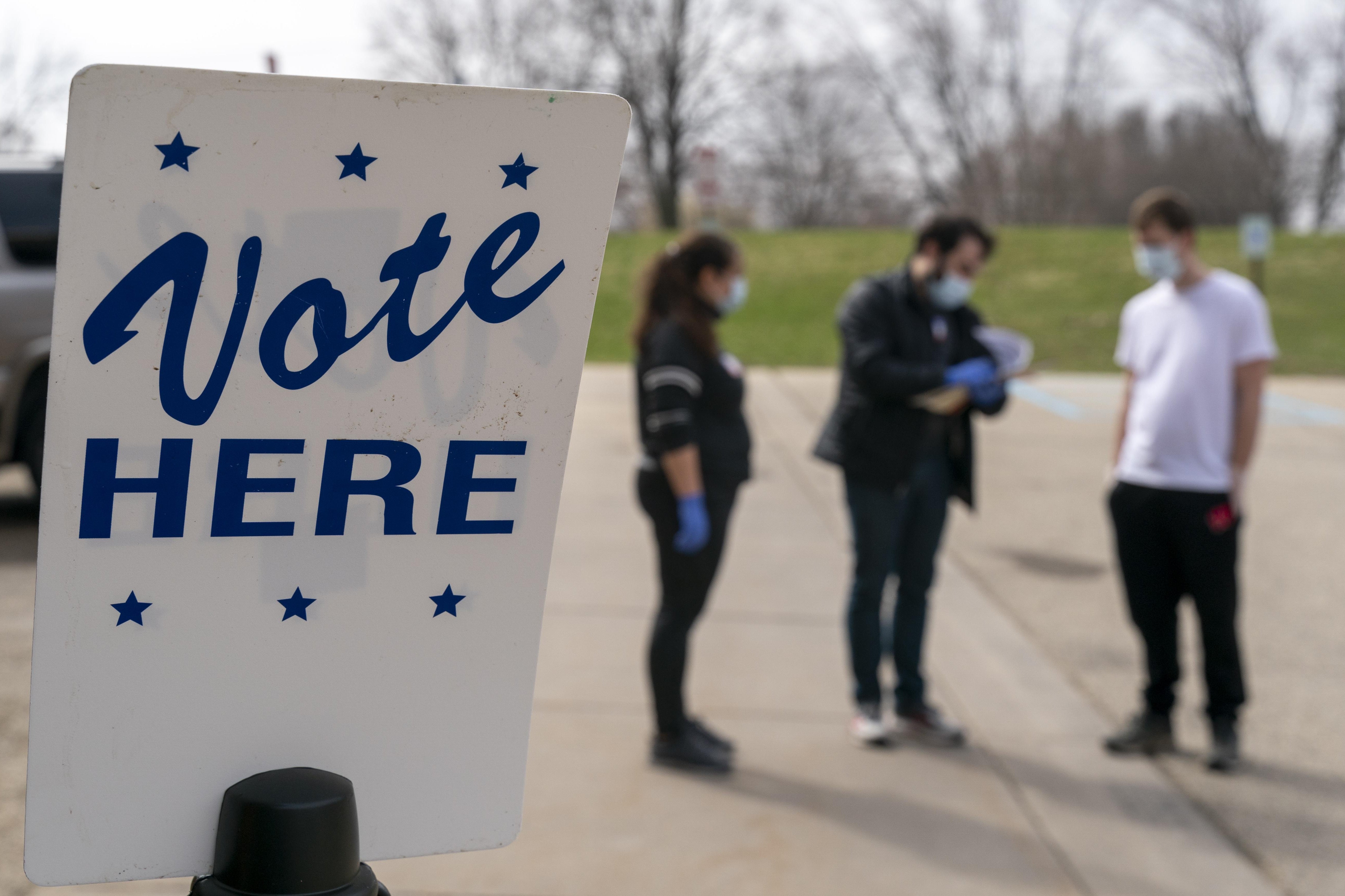 A "vote here" sign in front of three people standing in front of a polling place in Madison, Wisconsin during the 2020 election.