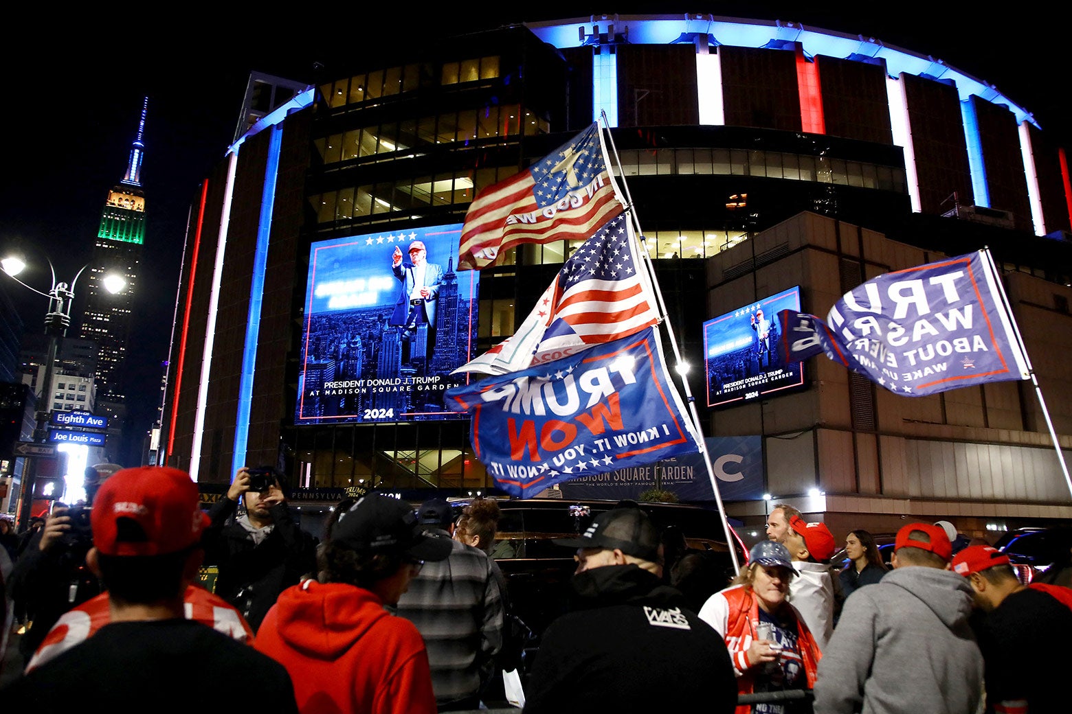 A crowd of people waving Trump flags outside a large arena featuring Trump campaign art.