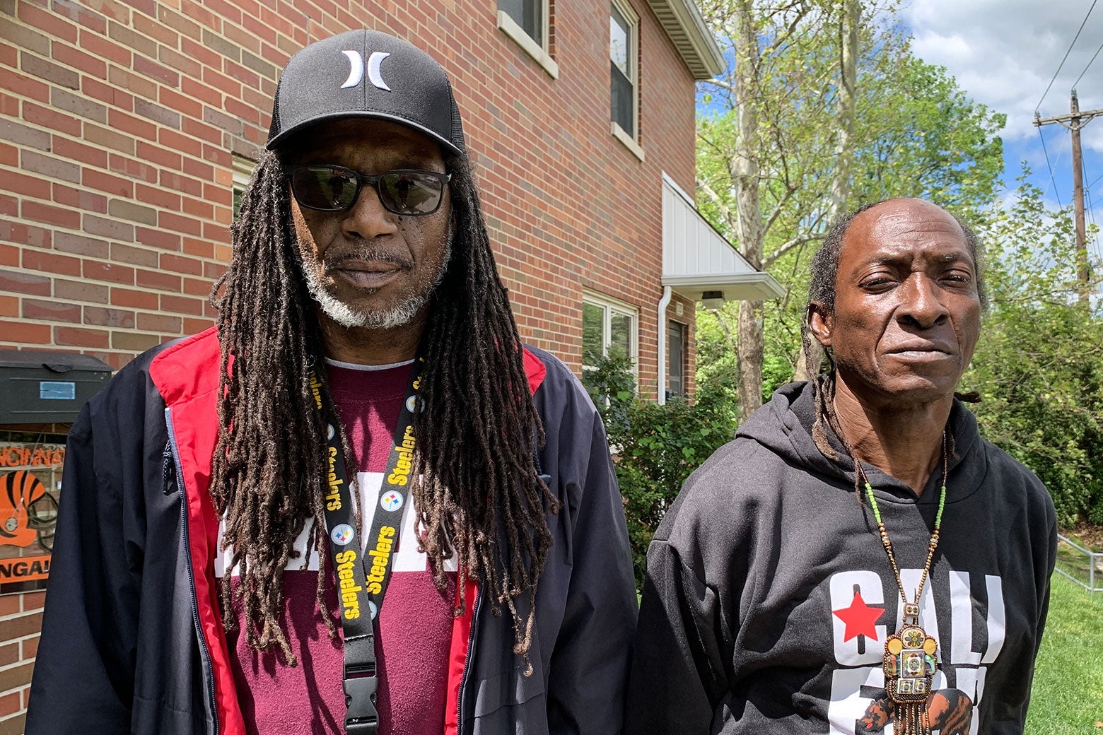Two Black men stand next to each other. One has long dreadlocks, sunglasses, and a Steelers lanyard. The other is wearing a colorful beaded necklace and a black sweatshirt.