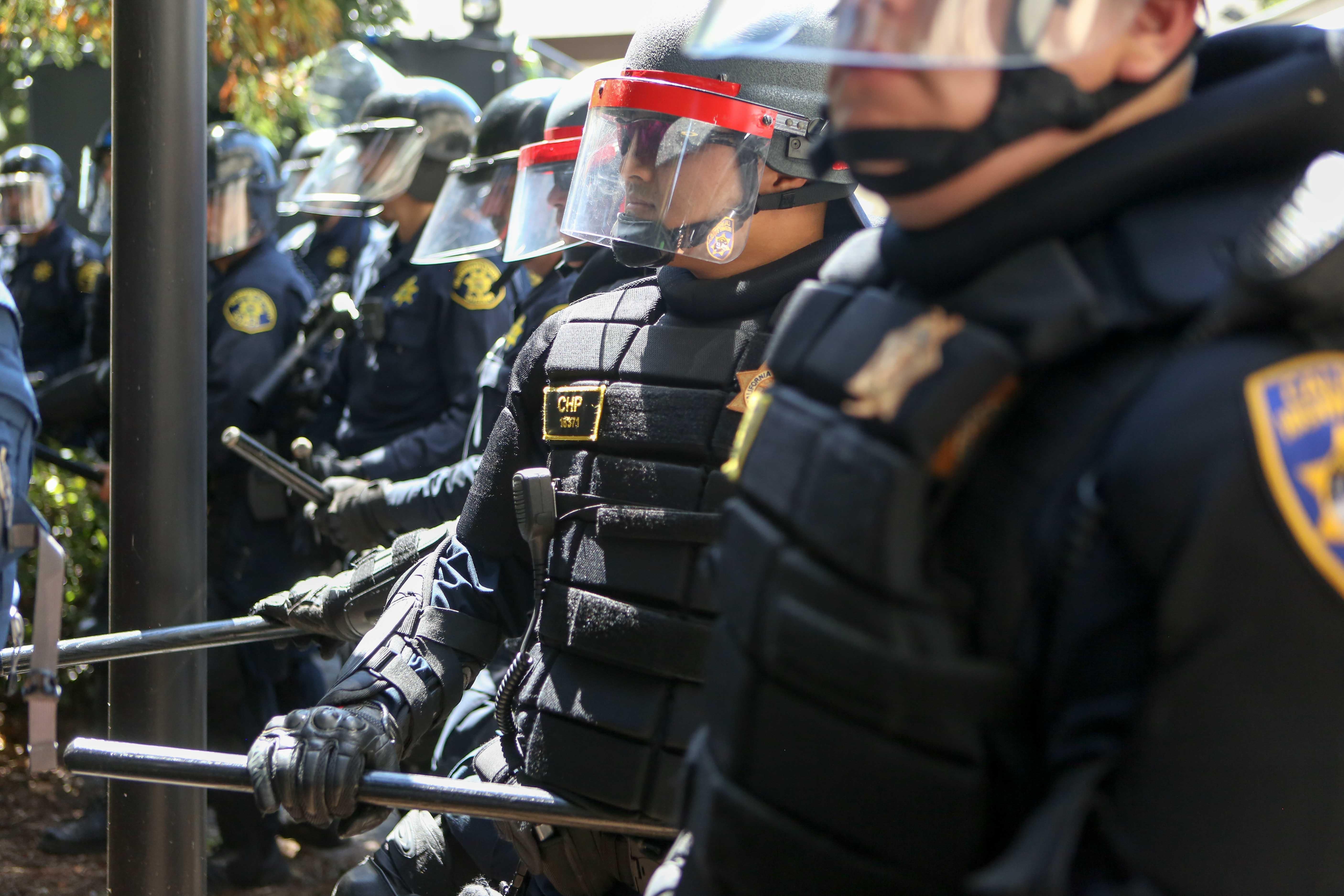 A row of police officers in heavy protective gear, some holding sticks.