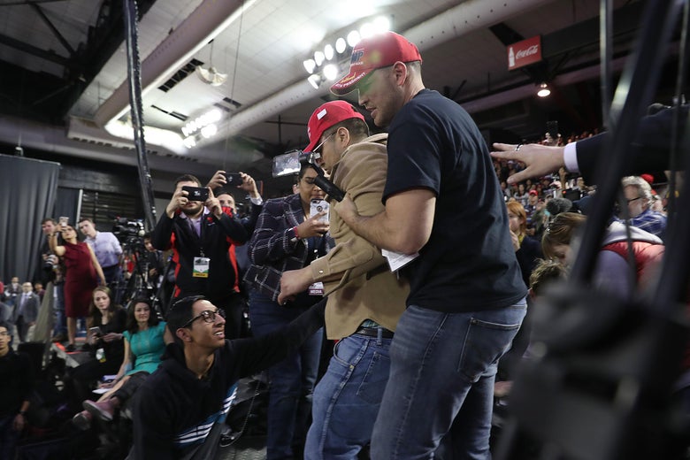 The man, wearing a brown shirt and red MAGA hat, is escorted off the platform, surrounded by reporters with cameras.