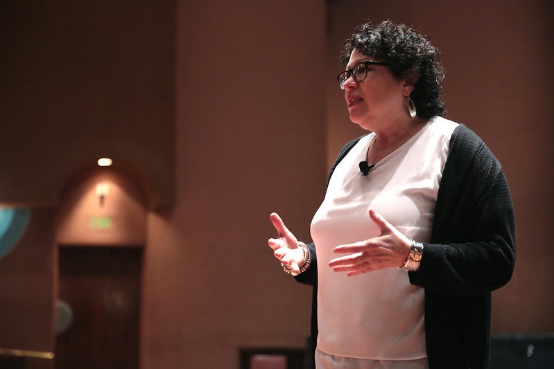 Sonia Sotomayor wearing a white shirt and a gray cardigan gestures as she speaks to a crowd