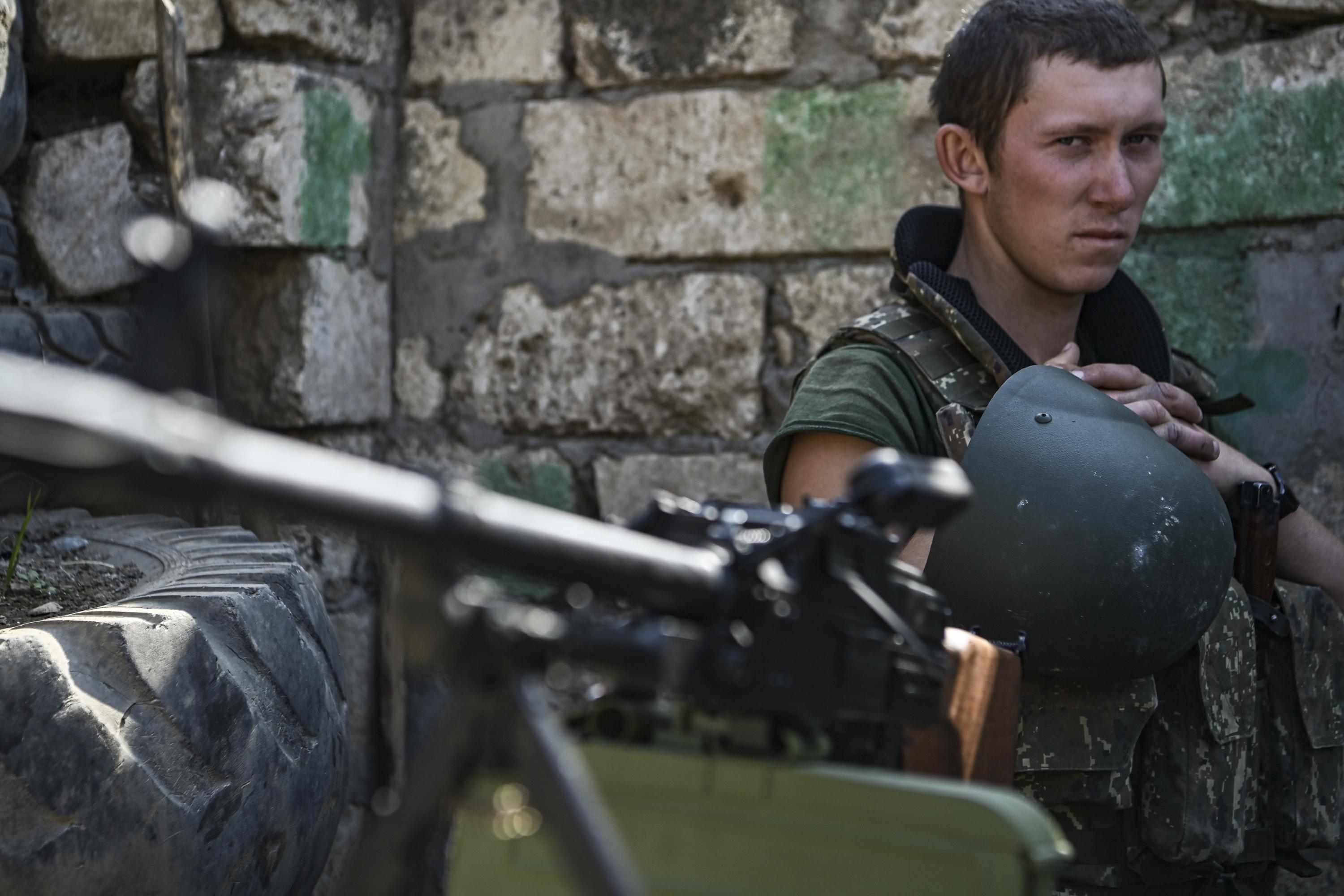 A young soldier with his helmet off looks over his rifle at the camera. 