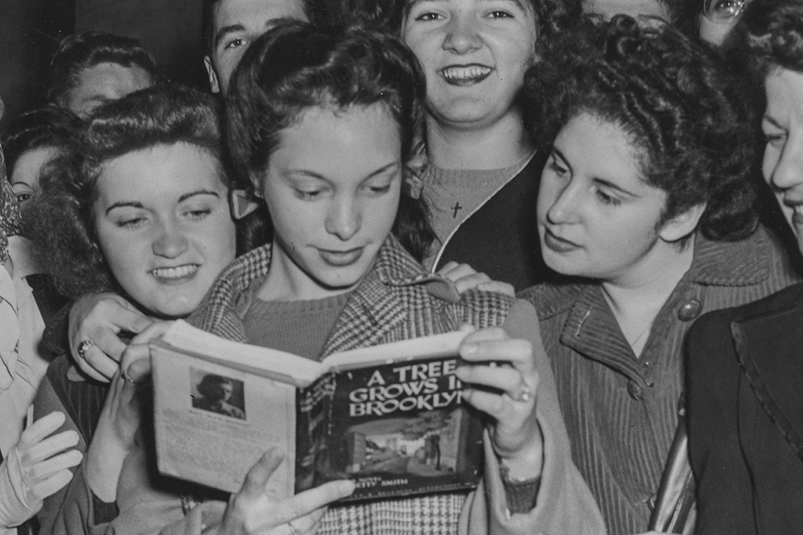 A group of young women in the 1940s smiling as they crowd around one reading A Tree Grows in Brooklyn. 