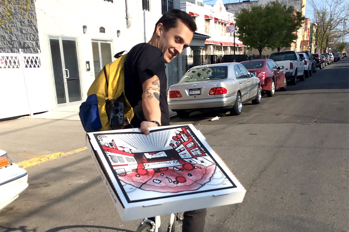 Liam Quigley riding a bicycle, looking backward and holding up a pizza box toward the camera.
