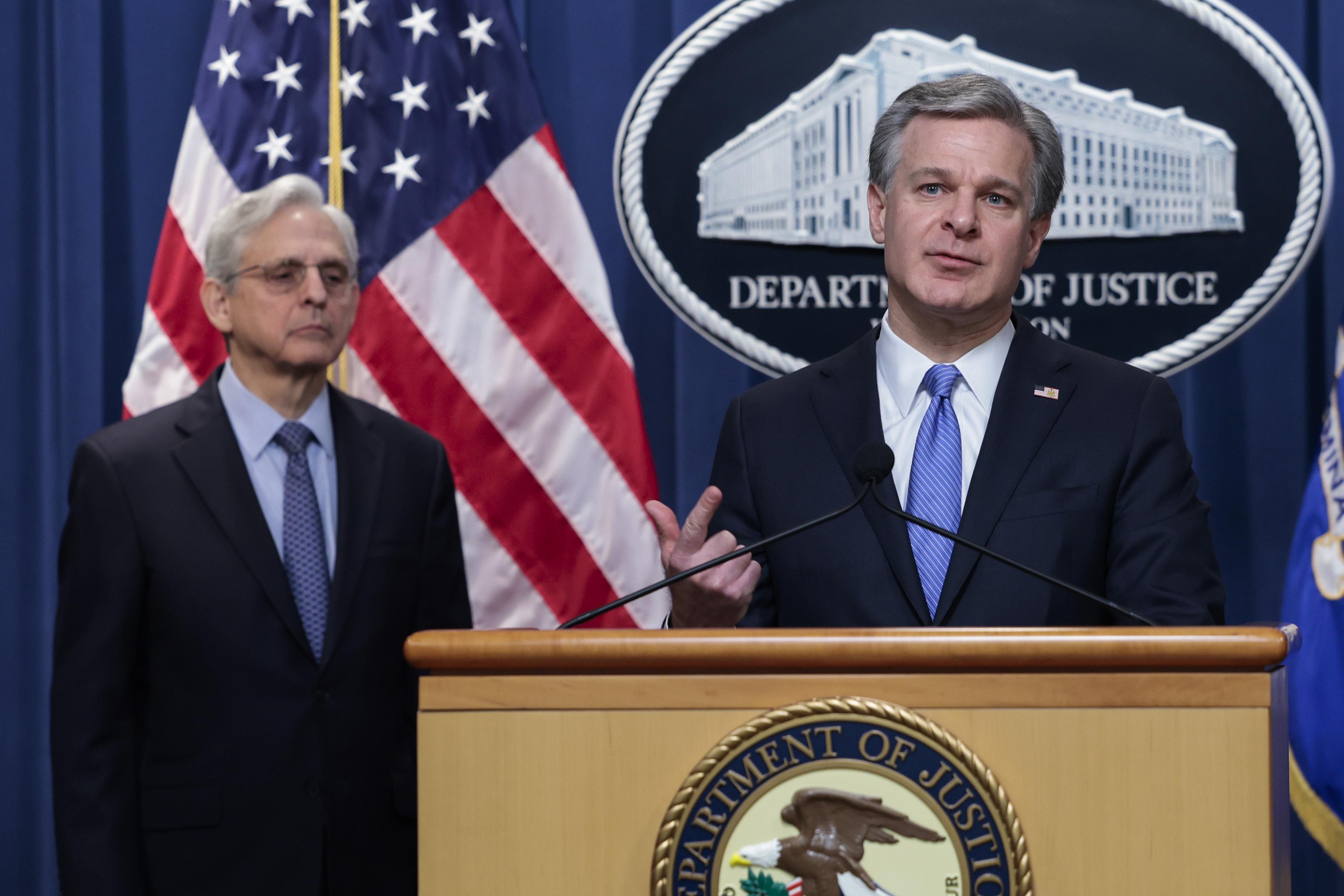 Wray points and speaks and Garland looks on. A Department of-Justice seal is on the podium in front of Wray, and curtain behind him.