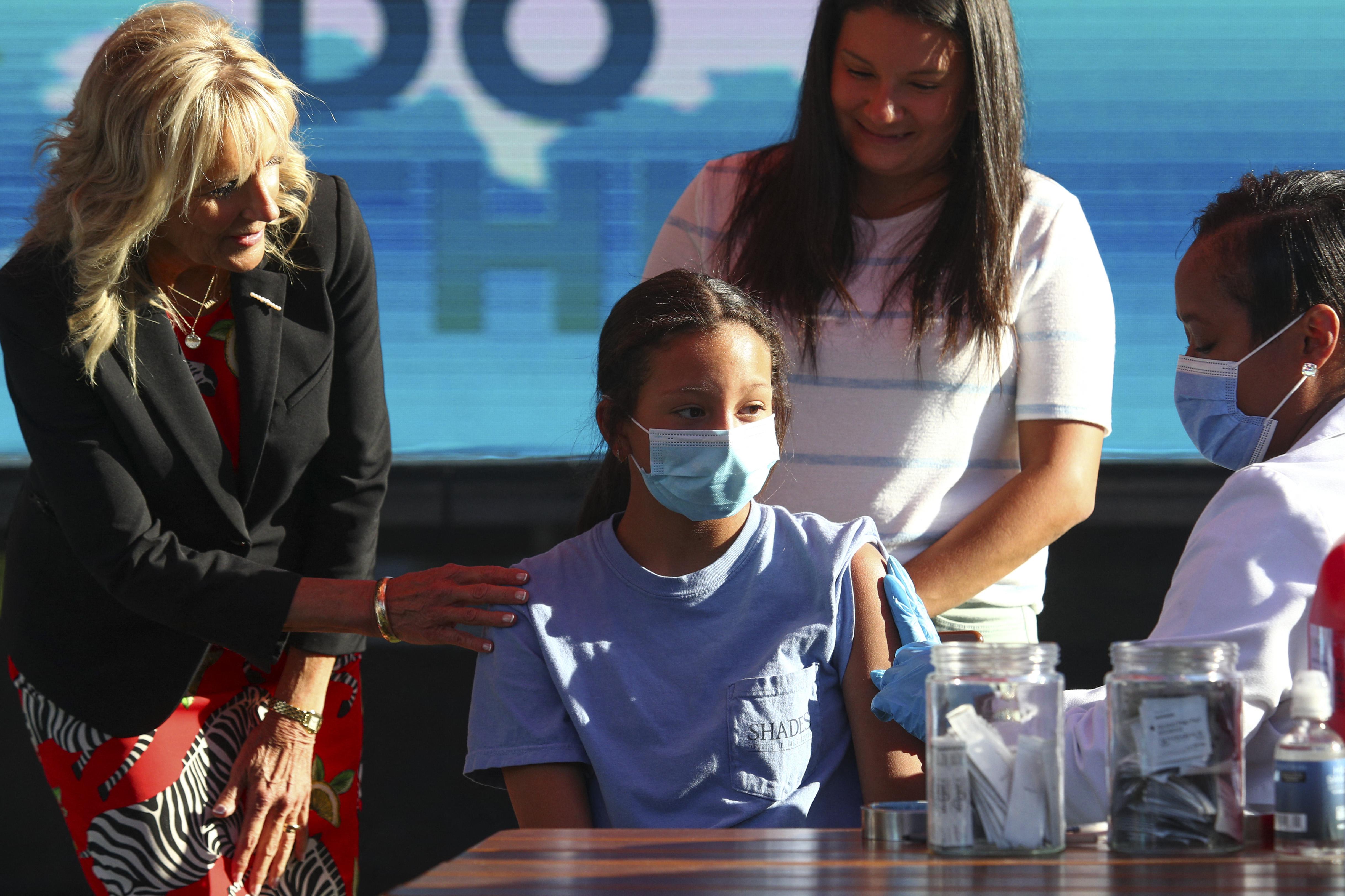 Jill Biden reaches an arm out to a 12-year-old girl wearing a mask who is receiving her COVID-19 vaccination. 