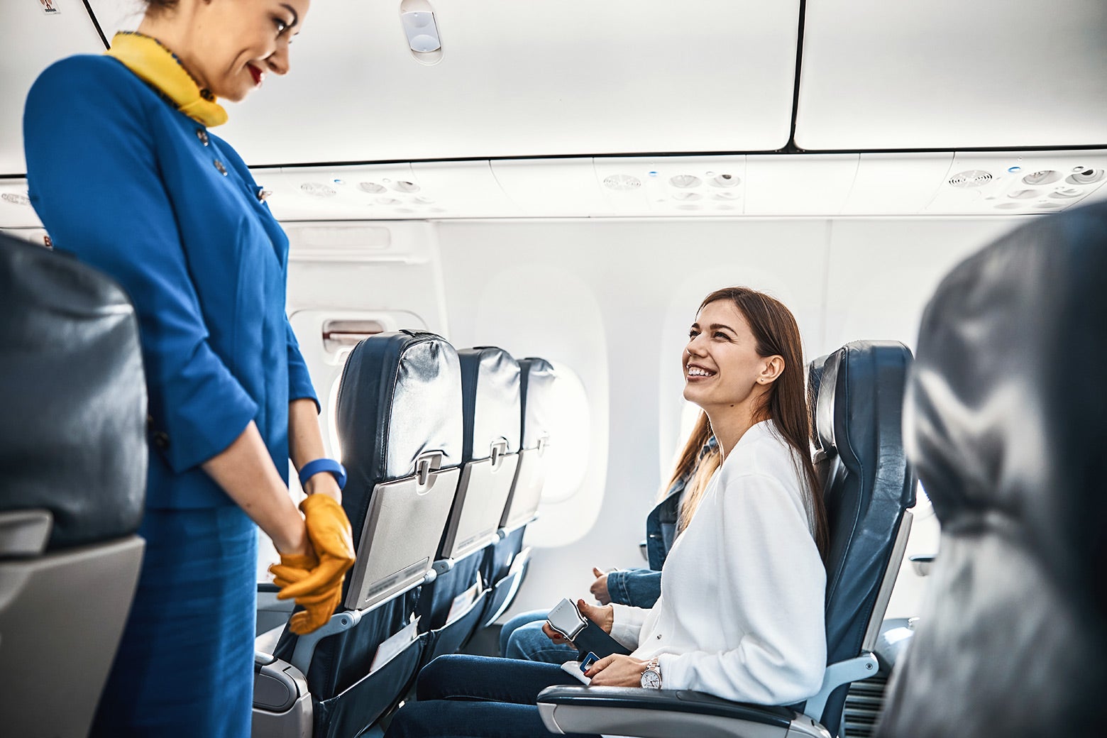 A woman seated in the aisle seat on a plane smiles at the blue-uniformed flight attendant. 