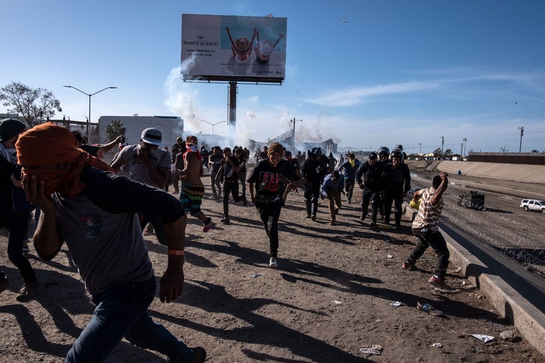 A crowd of migrants, many of them covering their mouths, flee from a cloud of gas behind them. 