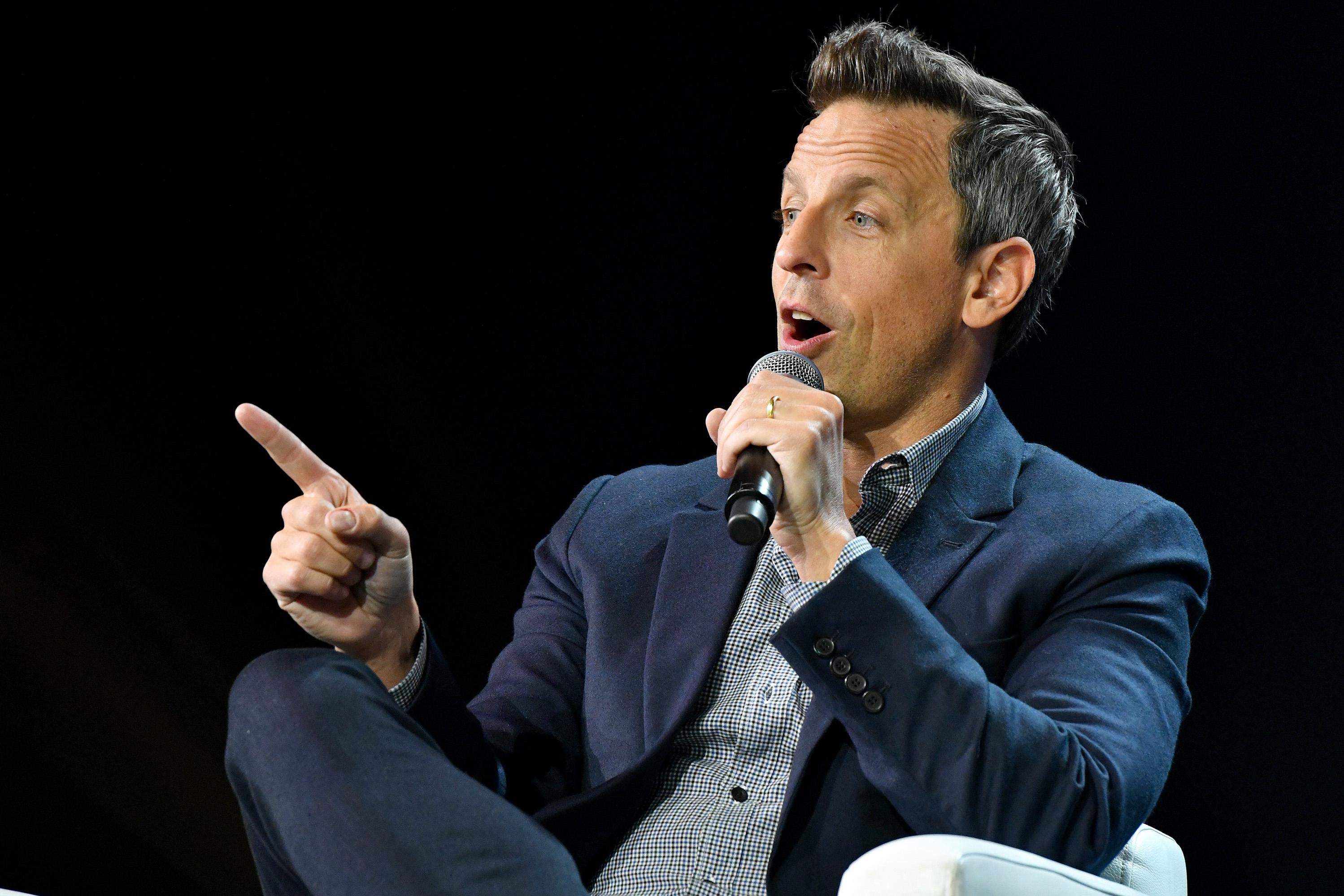 Seth Meyers, Host Late Night with Seth Meyers, holds a microphone and points his finger in front of a black backdrop.