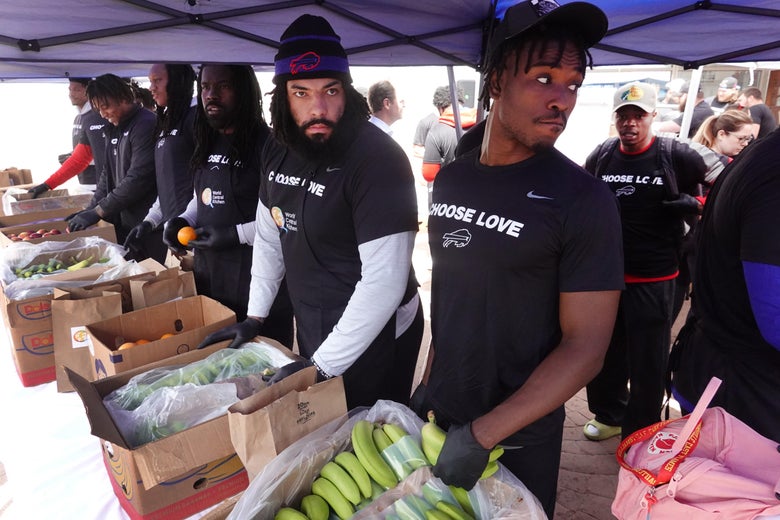 Football players wearing T-shirts that say "Choose Love" stand over cardboard boxes full of produce in a tent
