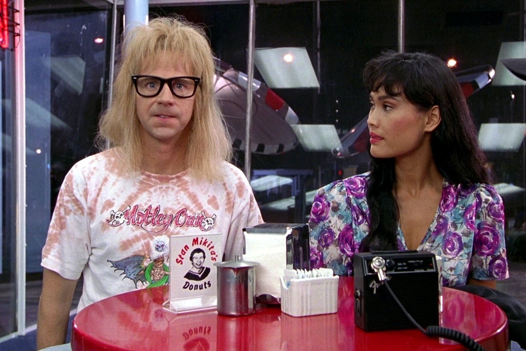 Dana Carvey, wearing thick, black-framed glasses, sits with Tia Carrere at a table. A small sign reads "Stan Mikita's Donuts."