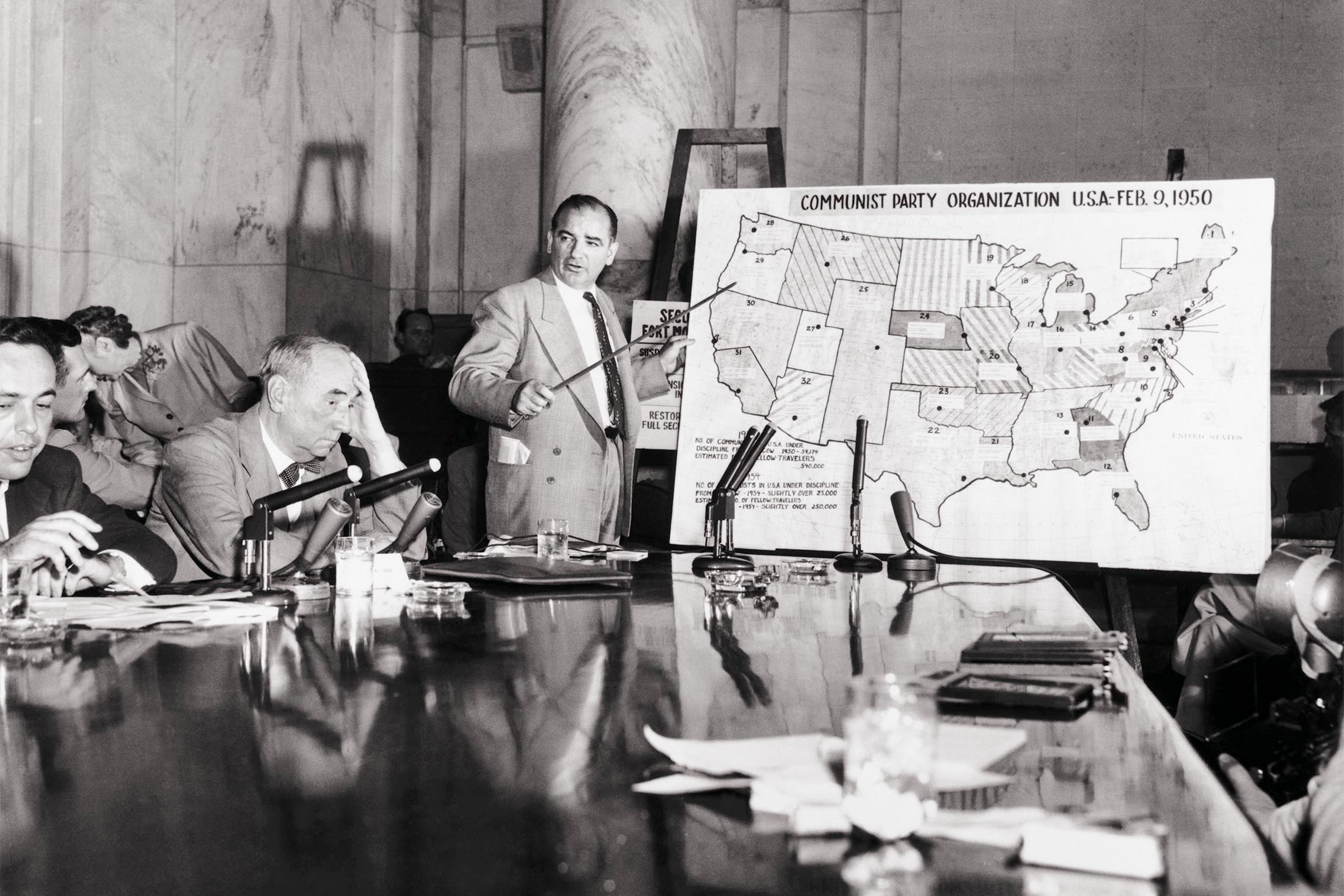 Sen. Joseph McCarthy points to a map depicting Communist Party organization across the U.S., in front of a conference table where Army counsel Joseph Welch and others are seated. Welch holds his head with his left hand in frustration.