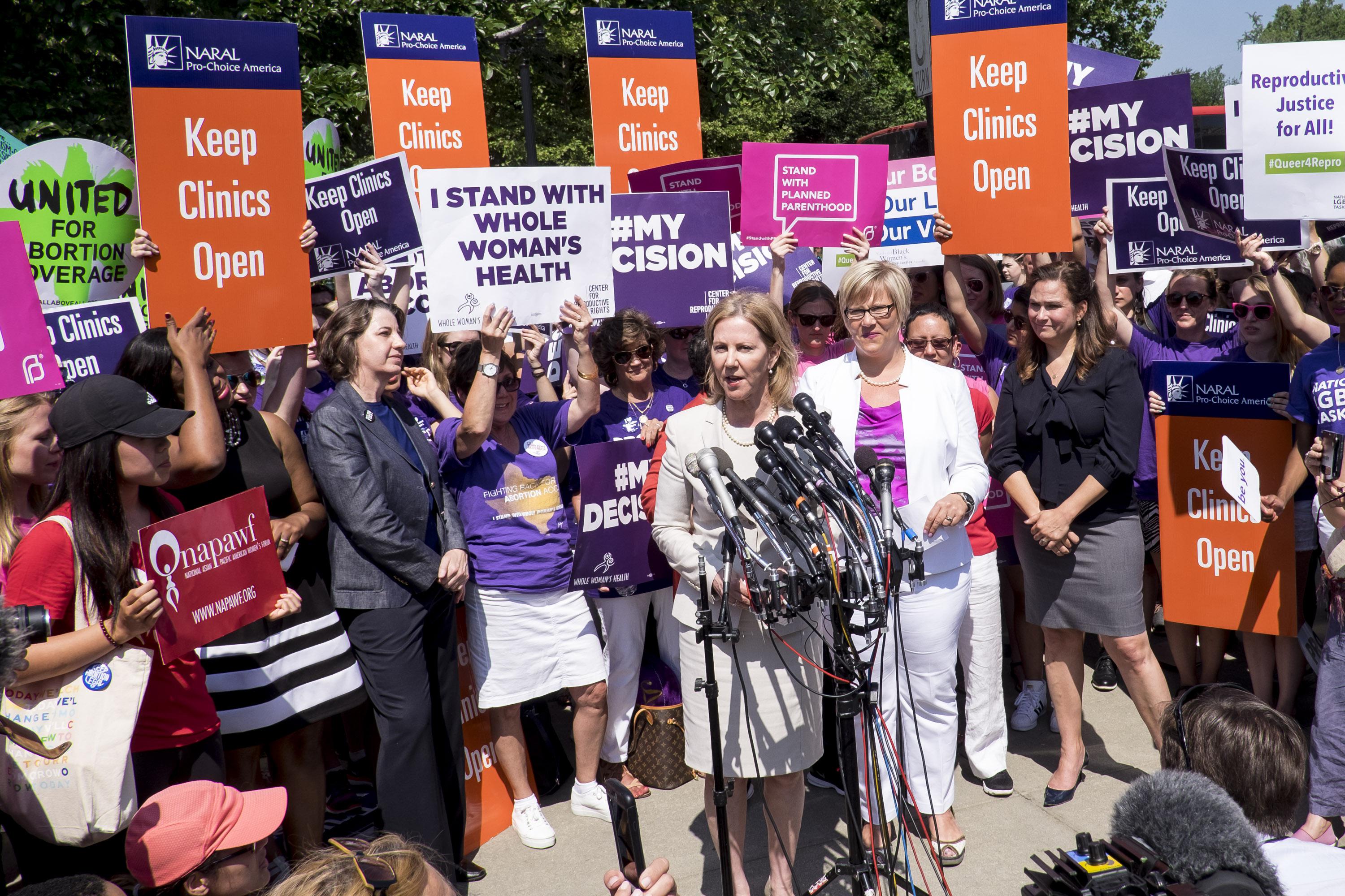 A crowd of people holding "Keep Clinics Open" signs crowd around two women at a podium.