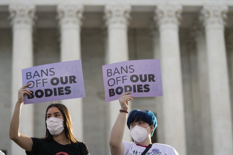 Two people wearing masks hold up signs that say "Bans Off Our Bodies" in front of the columns of the Supreme Court