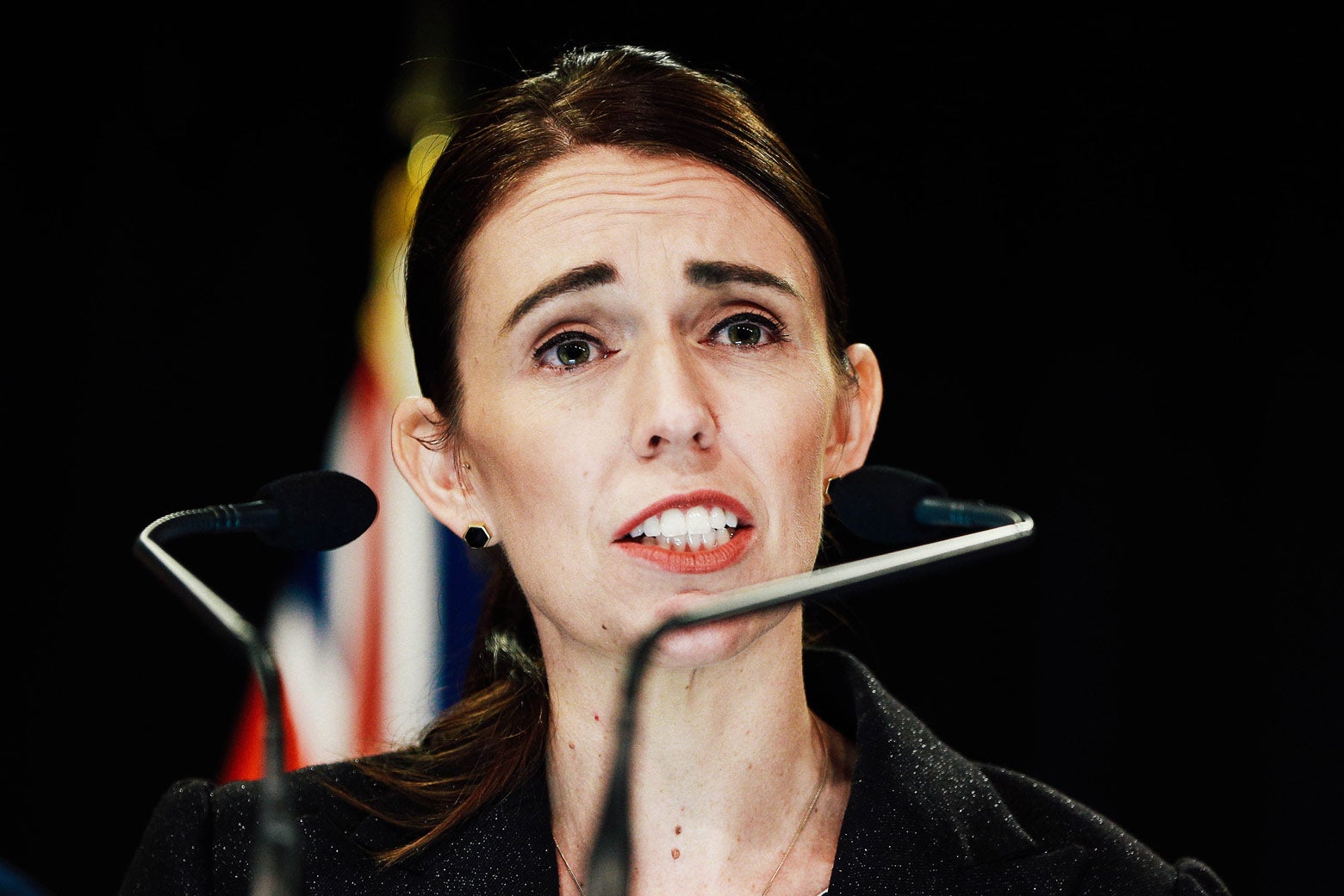 Jacinda Ardern behind microphones with a New Zealand flag in the background.