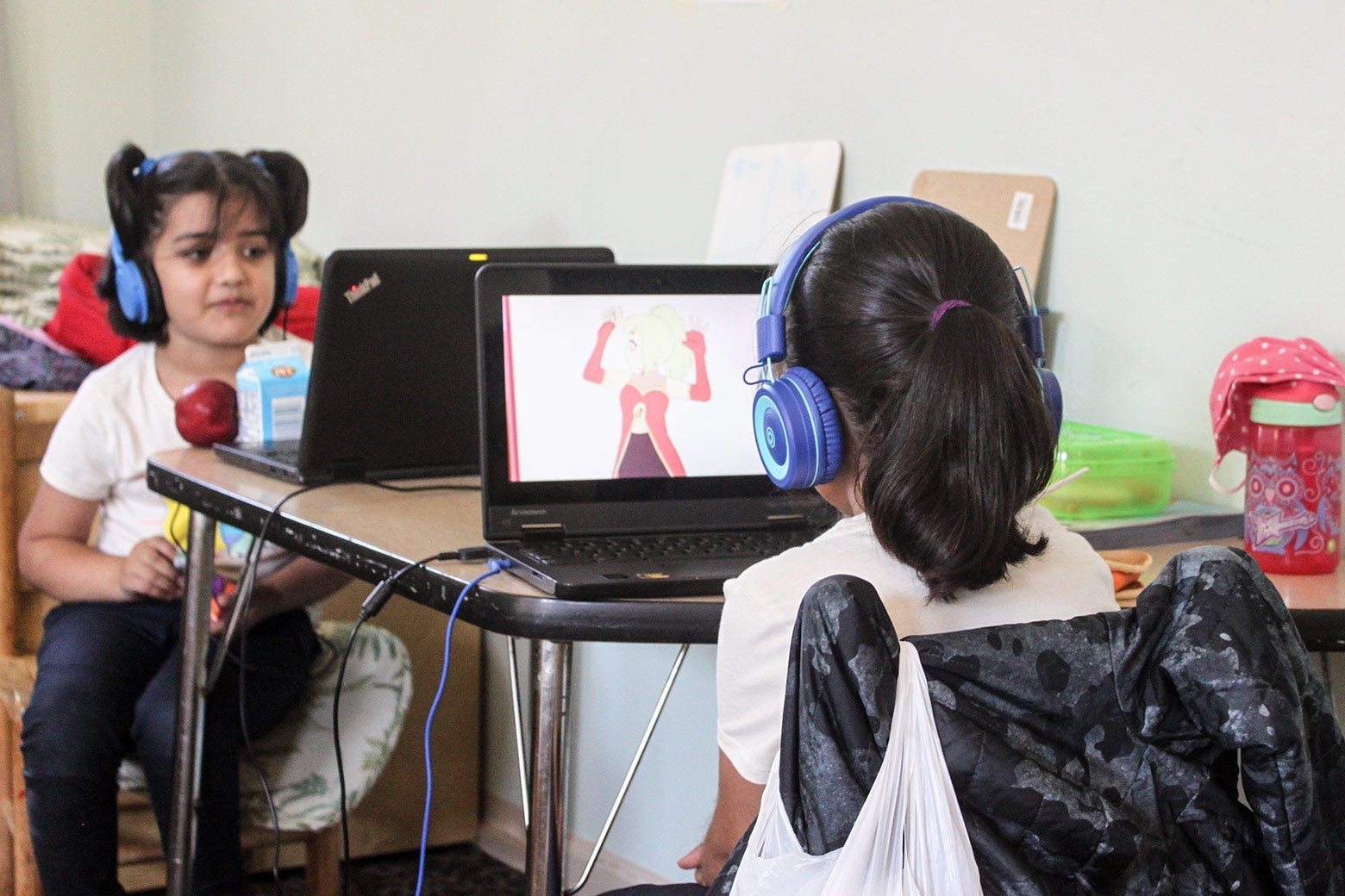 Two small children in large blue headphones sit behind laptops.