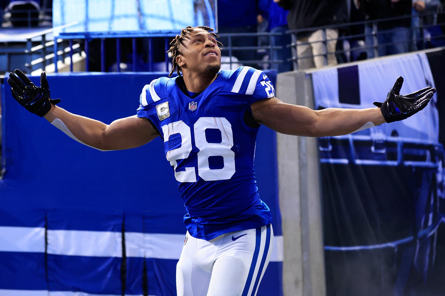 Taylor in pads and uniform with his helmet off looks back over his shoulder at fans with his arms outspread, encouraging them to cheer louder, as he runs onto the field.