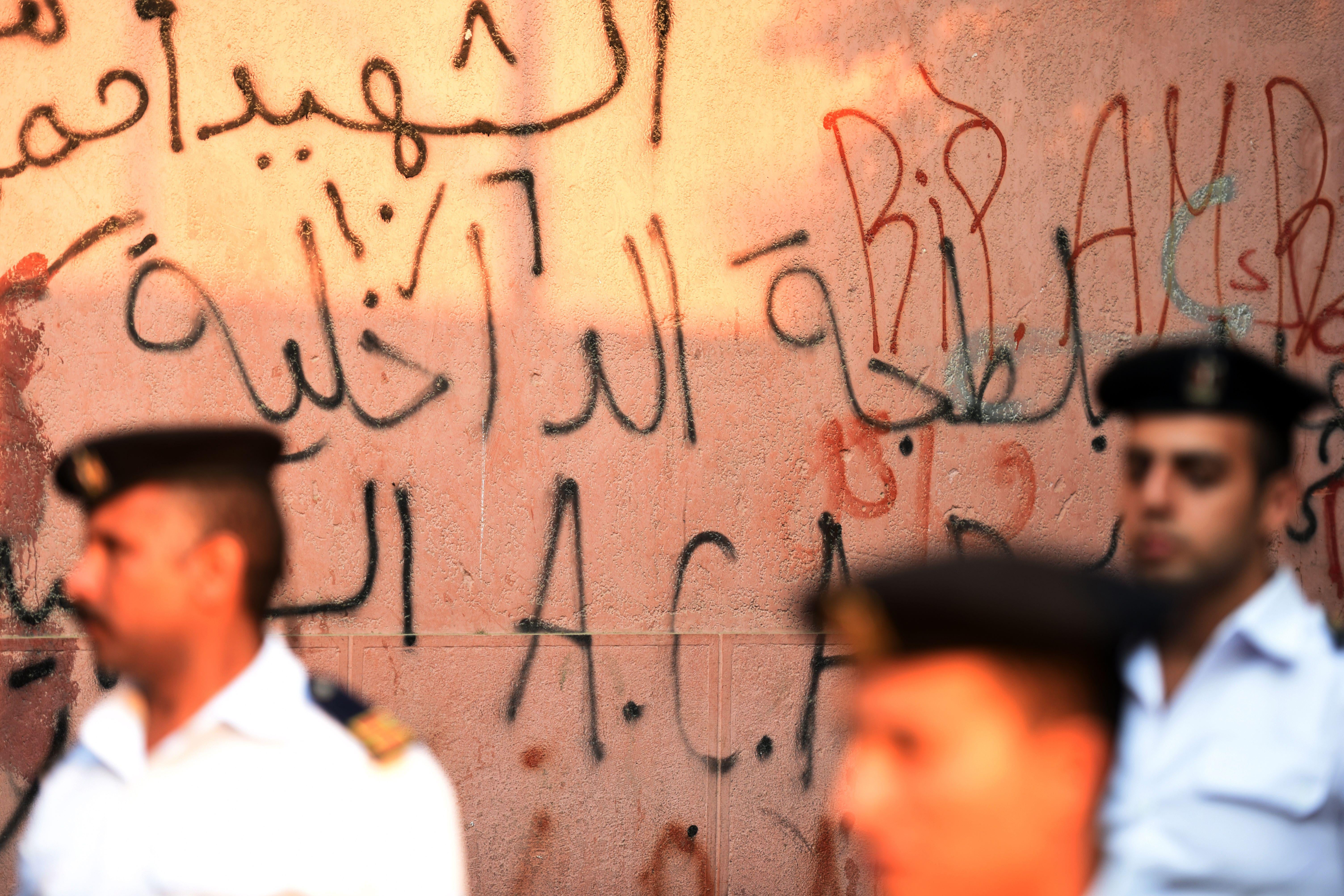 Egyptian police officers stand before a wall covered in graffiti.