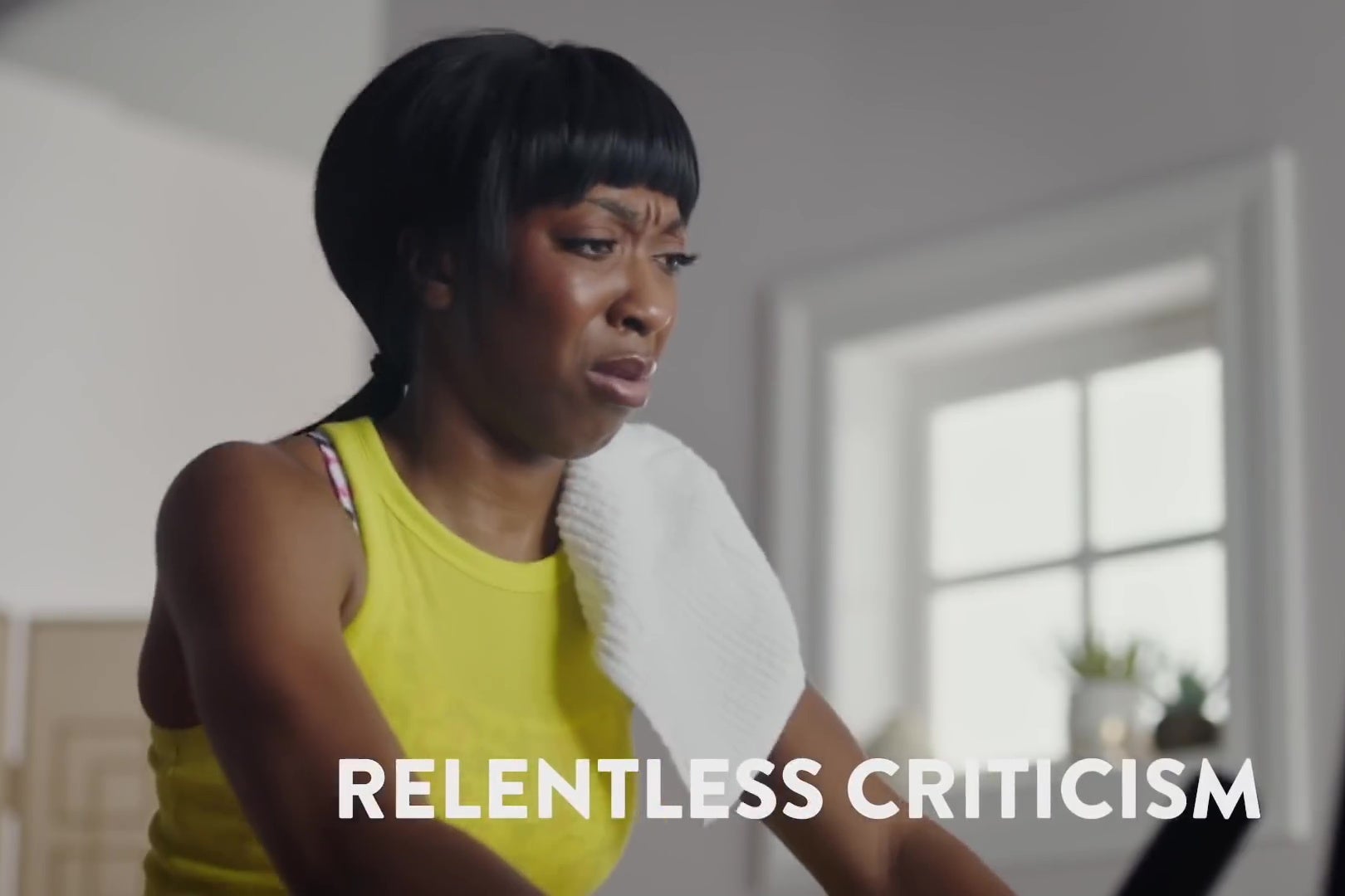 Ego Nwodim, on an exercise bike, looks at the view screen in disgust. A chyron reads "RELENTLESS CRITICISM."
