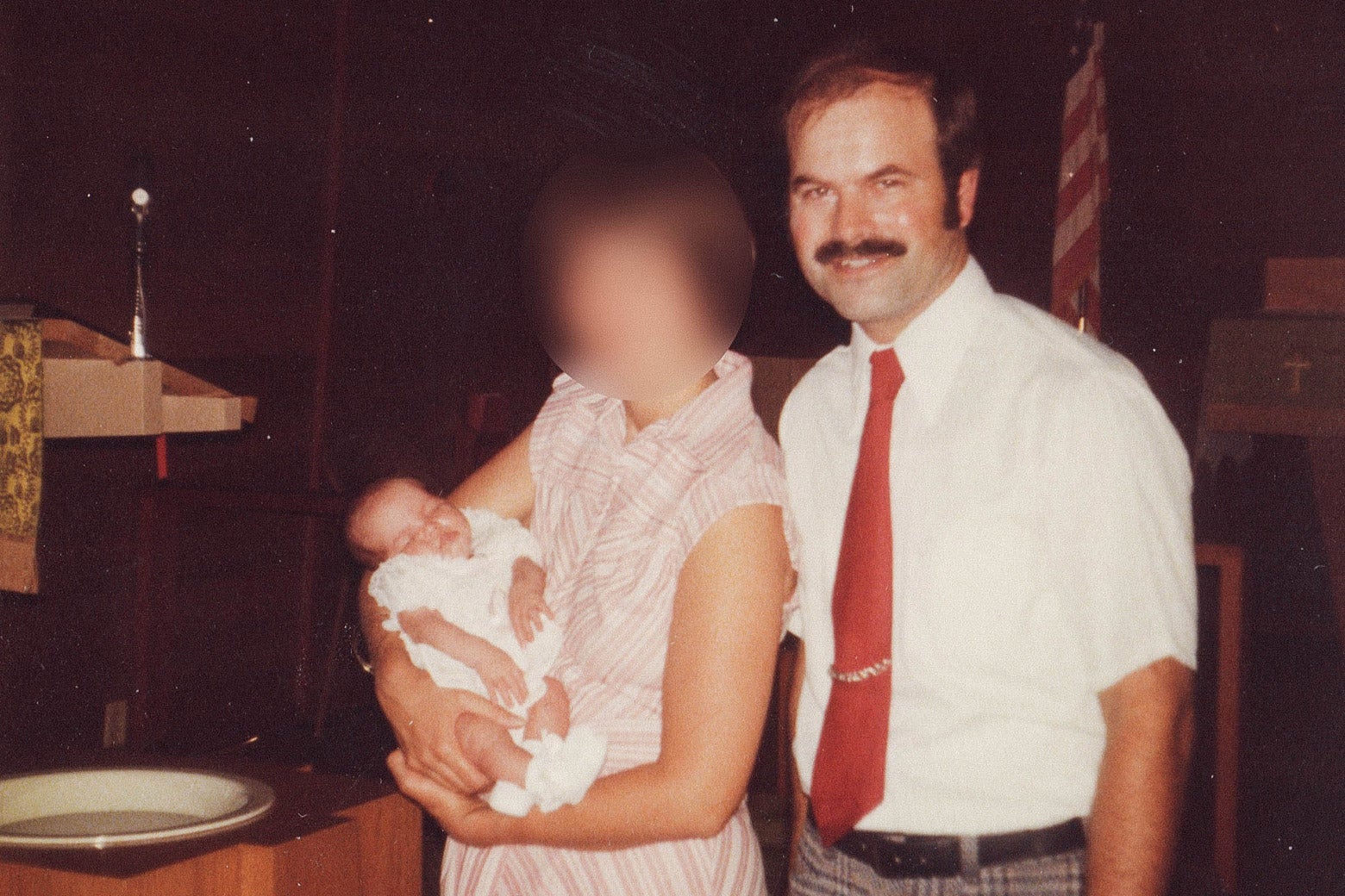 A woman whose face is blurred out holds a baby as the father stands beside her.