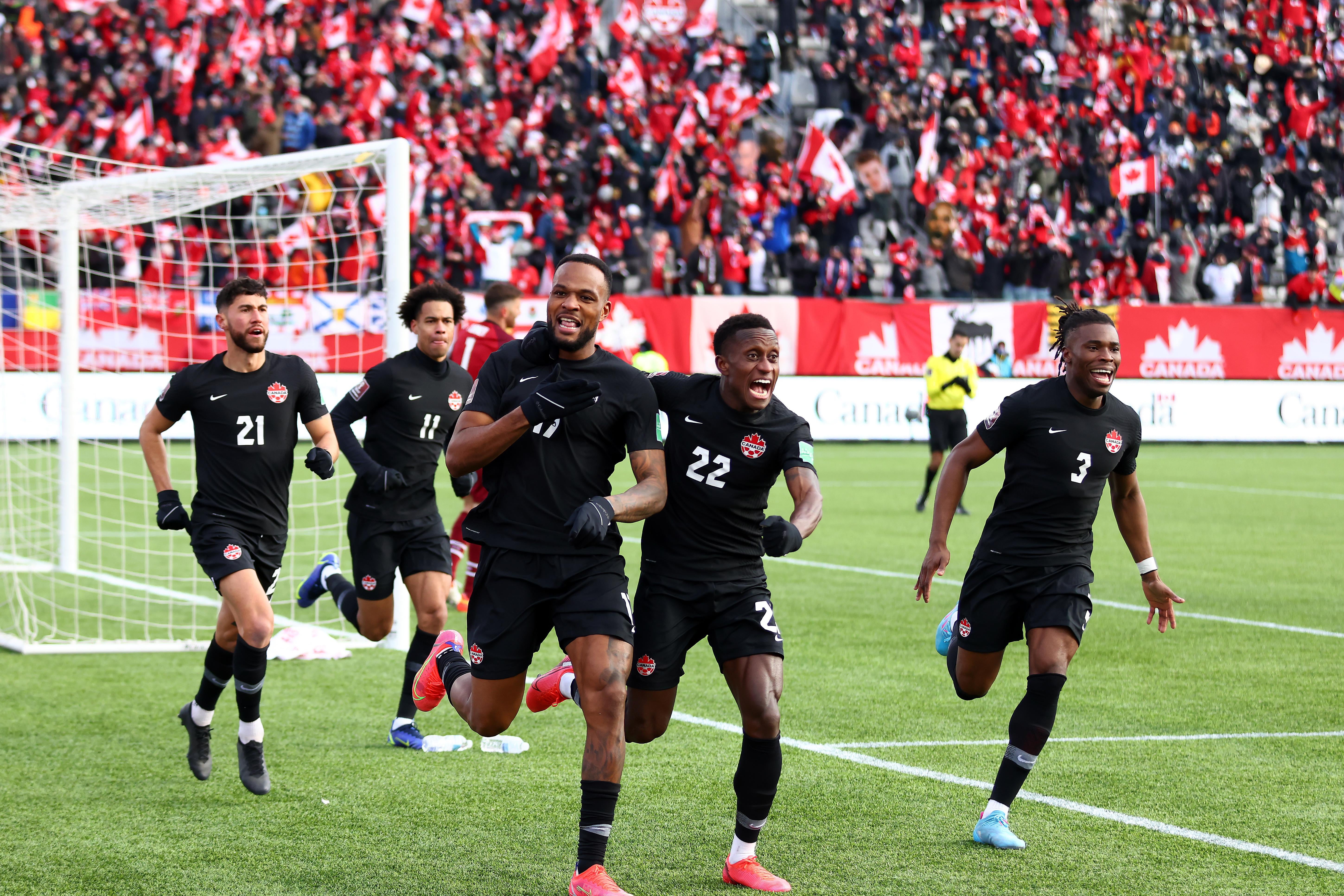 Larin, Laryea, and four other Canadian players run and cheer in front of the net, with the American goalie and ref on the field and Canadian fans waving maple leaf flags in the stands behind them