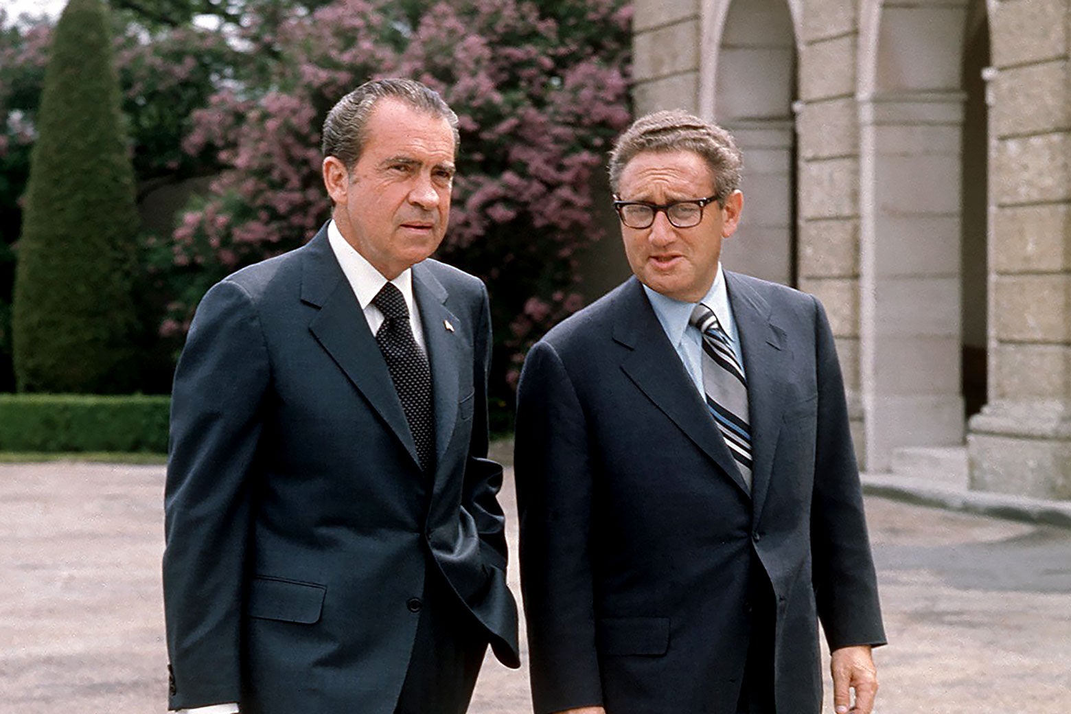Two middle-aged white men in suits walk and talk.
