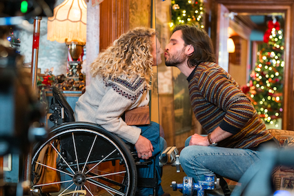 Actress Ali Stroker and her costar Daniel di Tomasso kiss the plexiglass between them. Christmas decorations light up the room behind the two of them.