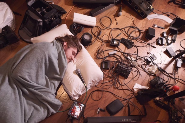 An overhead shot of comedian Bo Burnham lying on a floor in
        a sleeping bag, surrounded by microphone cords, talking into a
        microphone.