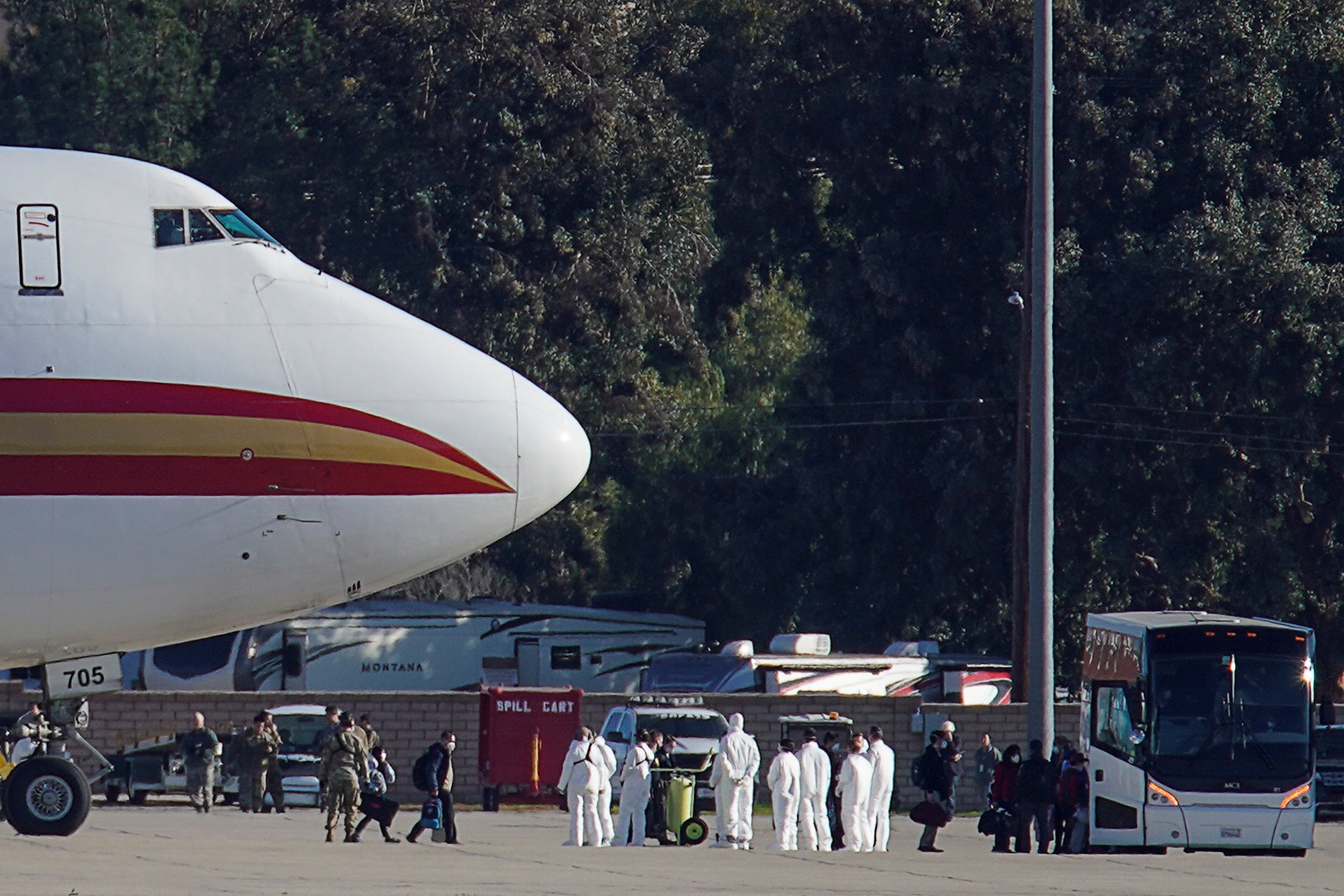 Passengers walk away from a jet and towards a bus. A group of officials in white protective clothes stand between the jet and the bus. 