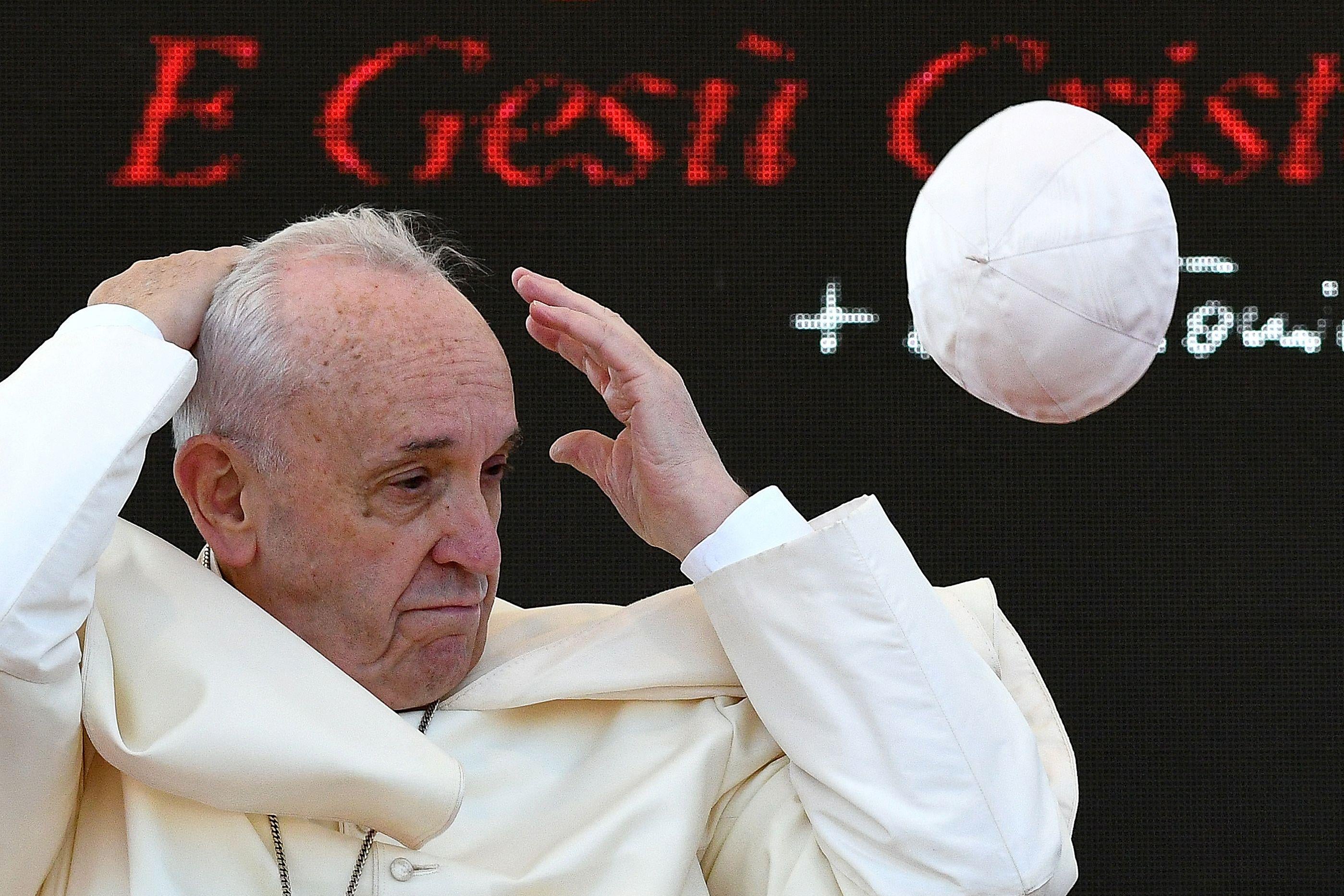 Pope Francis reaches his hands to his head as his zucchetto (skullcap) blows away in the wind.
