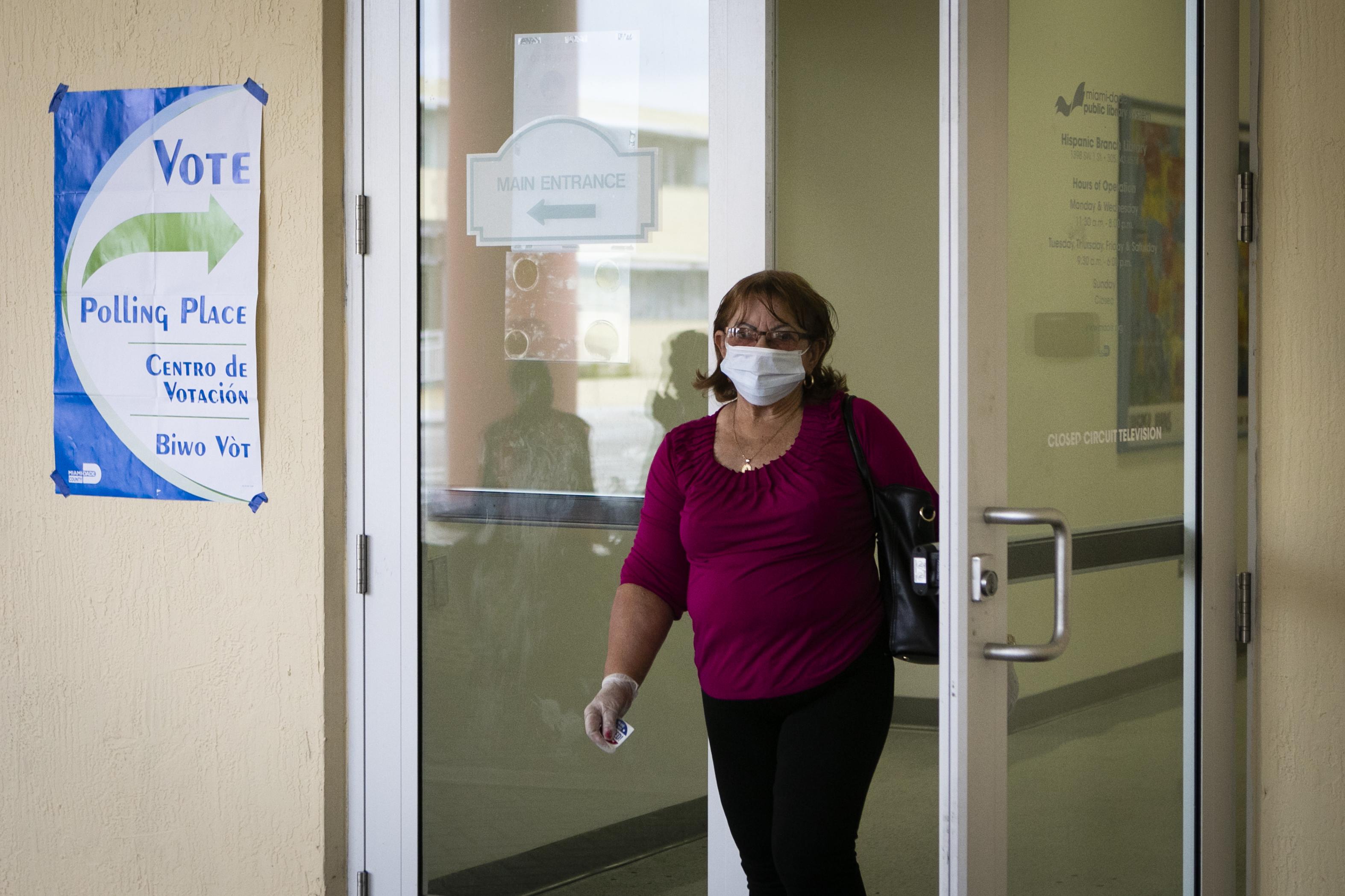 A woman wearing a surgical mask and gloves exits the polling place. A "VOTE" sign hangs on the wall next to the doors.