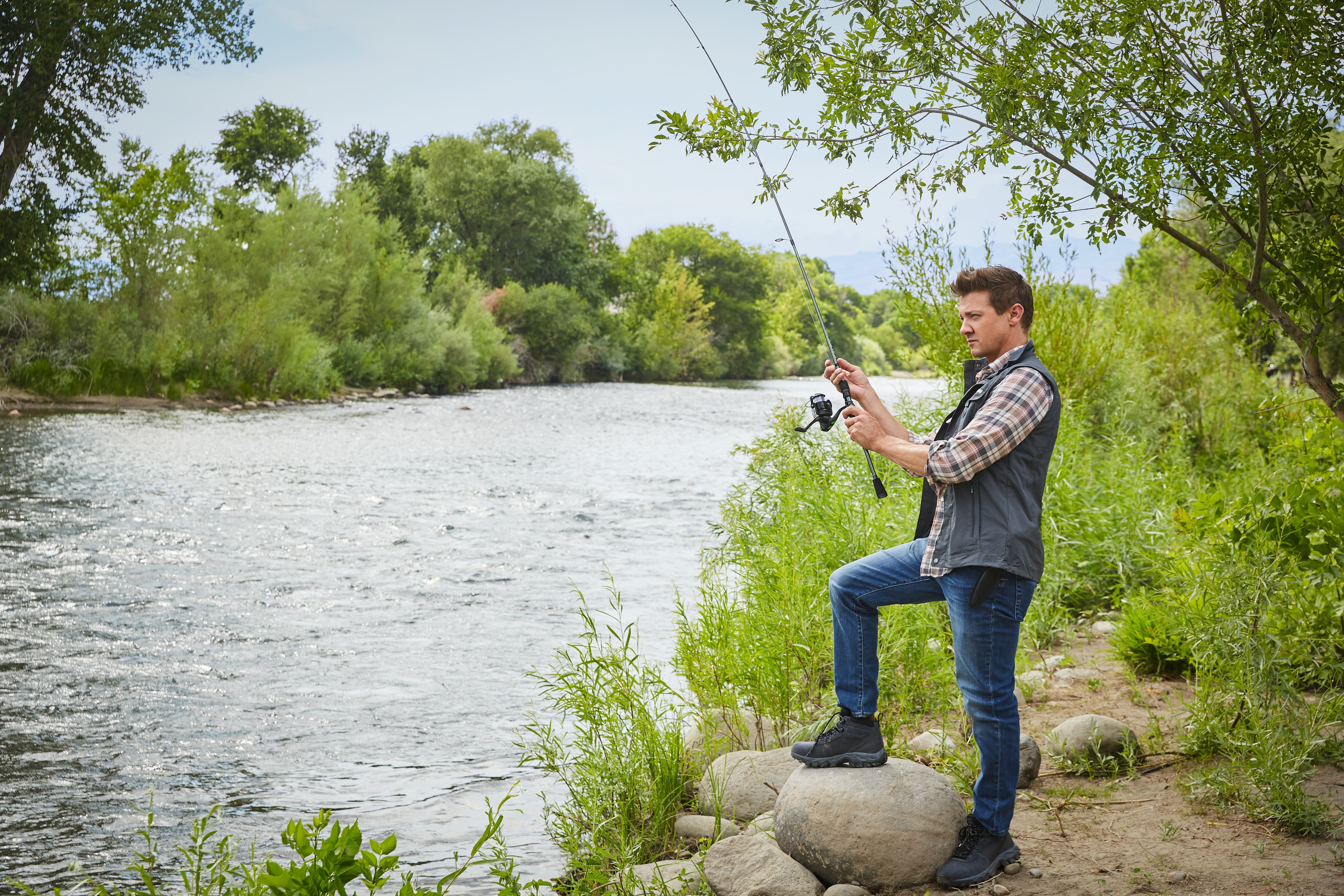 Jeremy Renner stands with one foot on a rock, holding a fishing rod that appears to have no line cast.