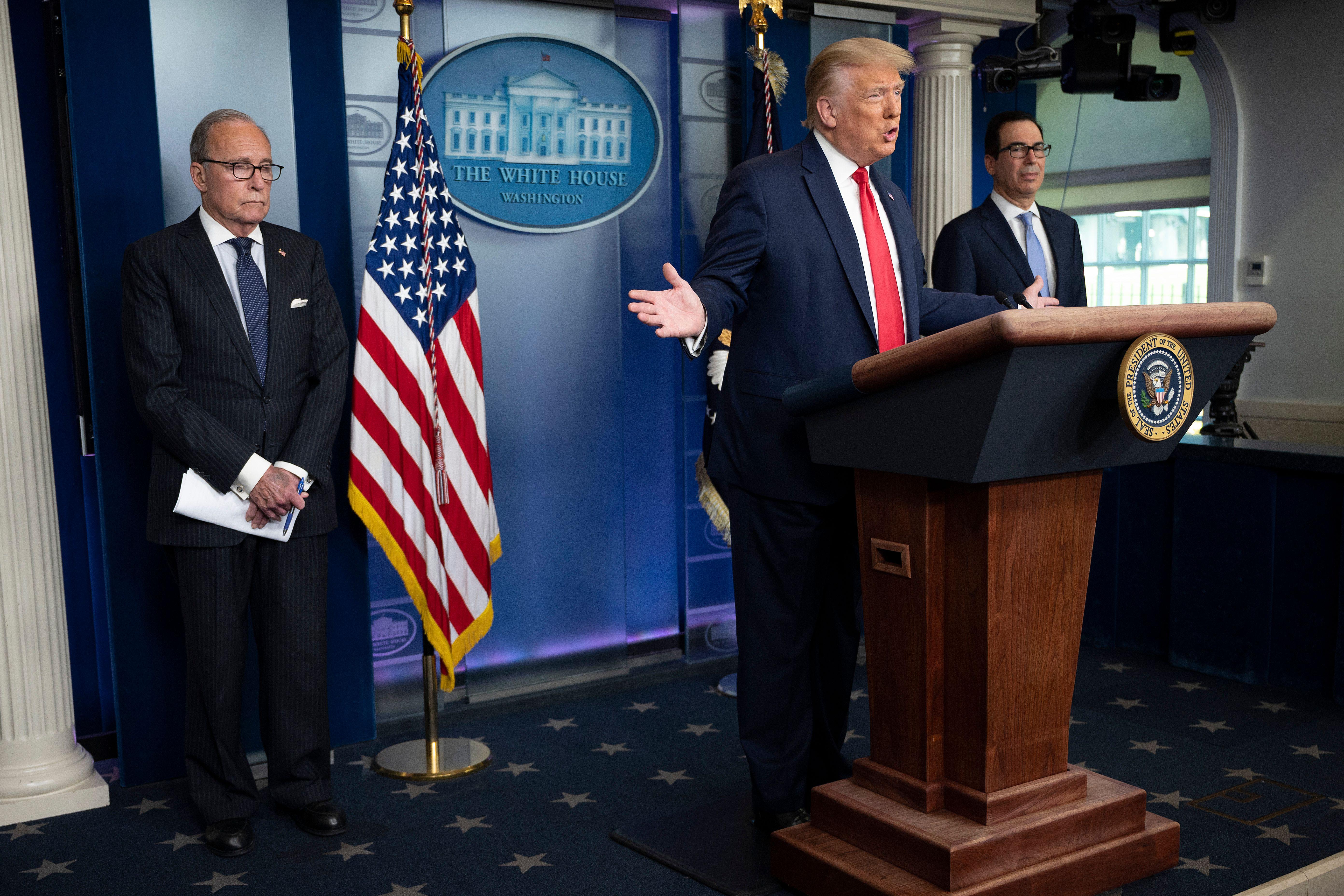 Donald Trump at the podium in the White House briefing room with his arms spread wide. Kudlow and Mnuchin stand at either side of him.