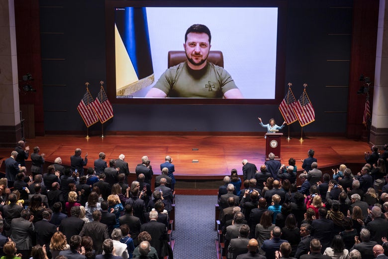 Zelensky's face is seen on a large screen displayed above a stage in a large auditorium with an audience.