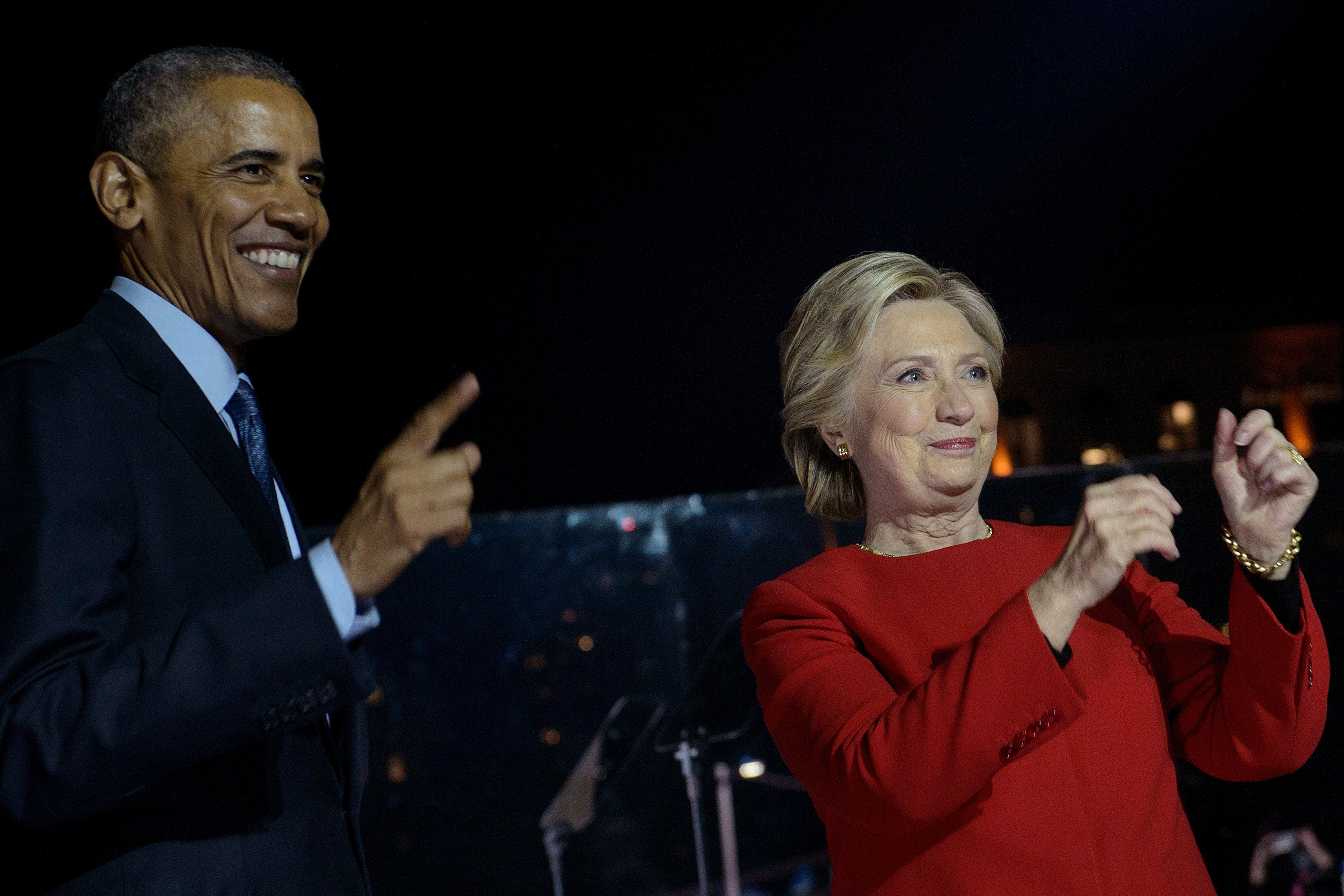 On a campaign rally stage, Obama smiles and points to the crowd while Hillary Clinton gestures with her hands.