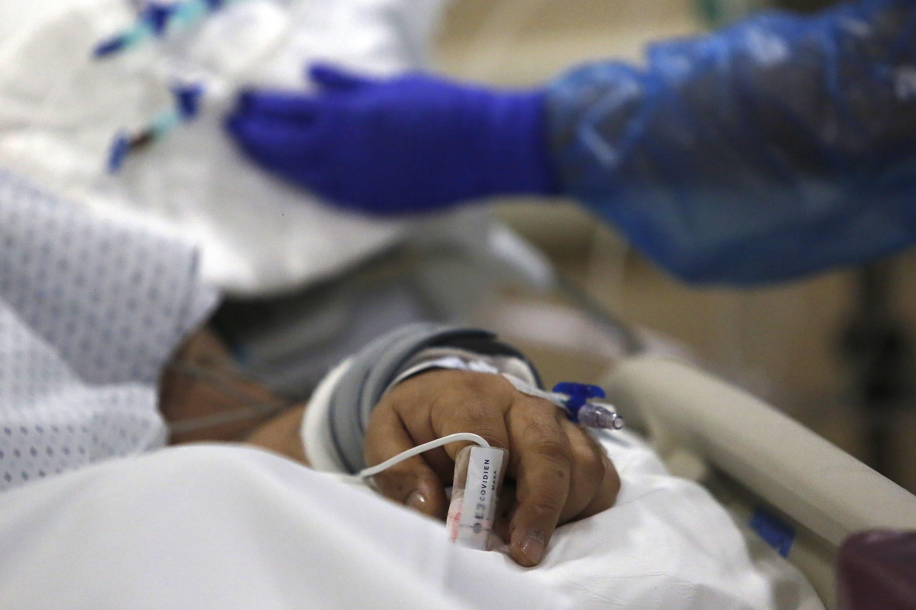 A close-up of a patient's hand on a hospital bed as gloved hands work nearby.
