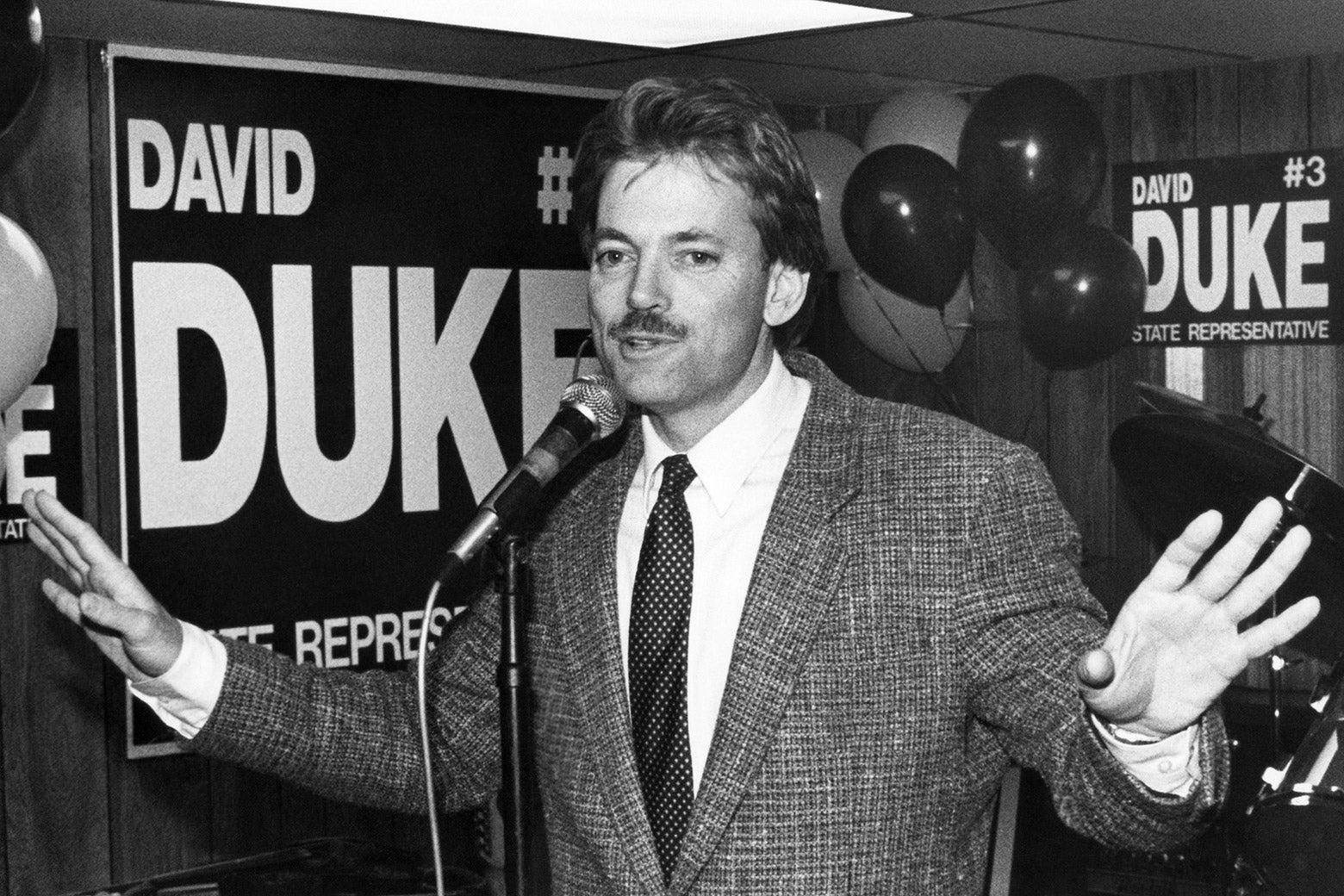 David Duke at a microphone in a suit and tie with campaign signs and balloons behind him