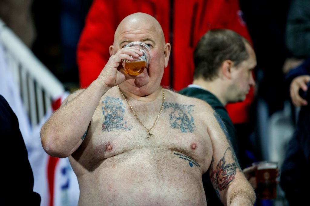 A large, shirtless, tattooed bald man drinks from a mostly empty clear cup of beer.