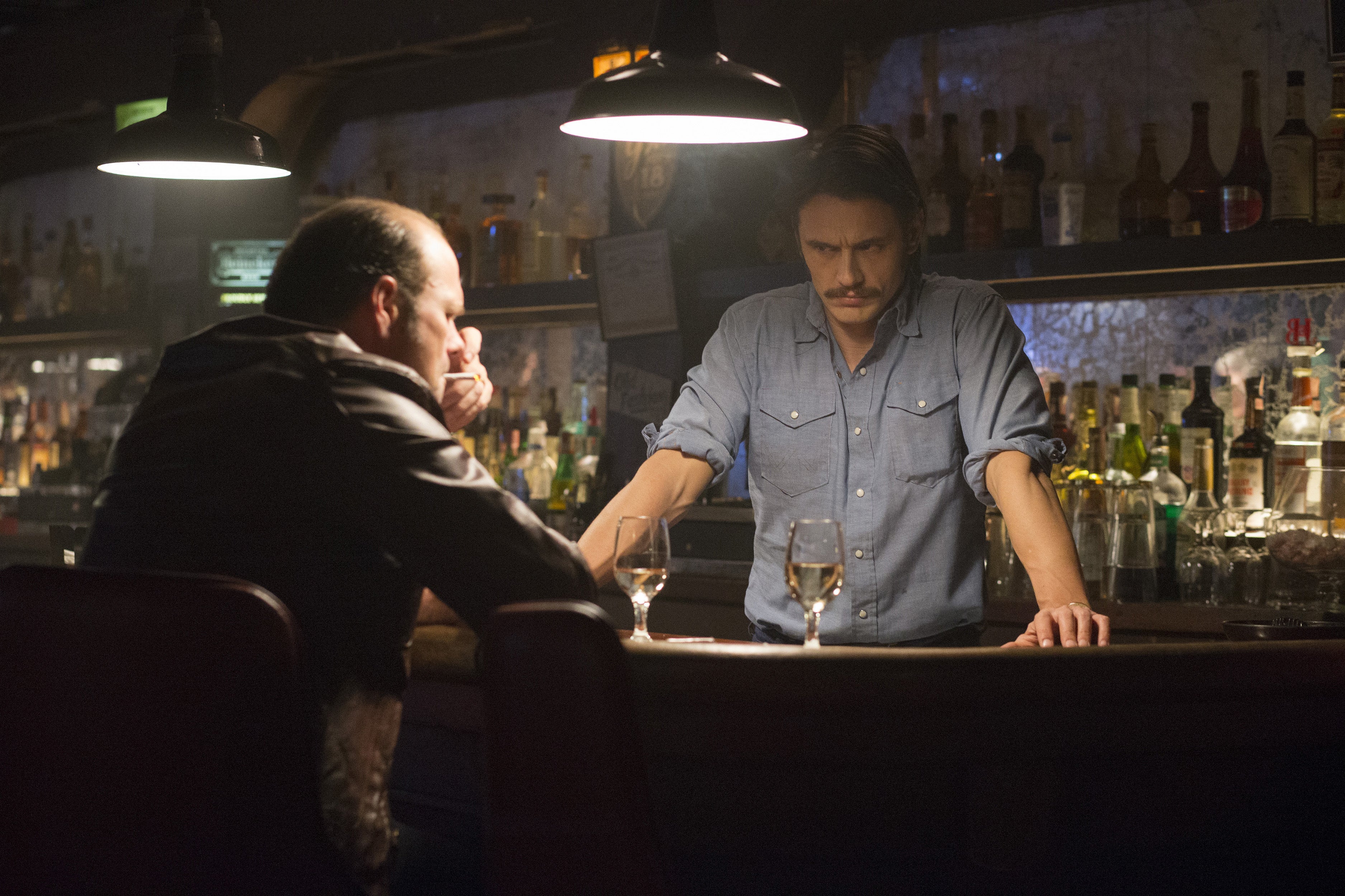 Chris Bauer sits at a bar, while James Franco, with a prominent mustache, stands behind it.