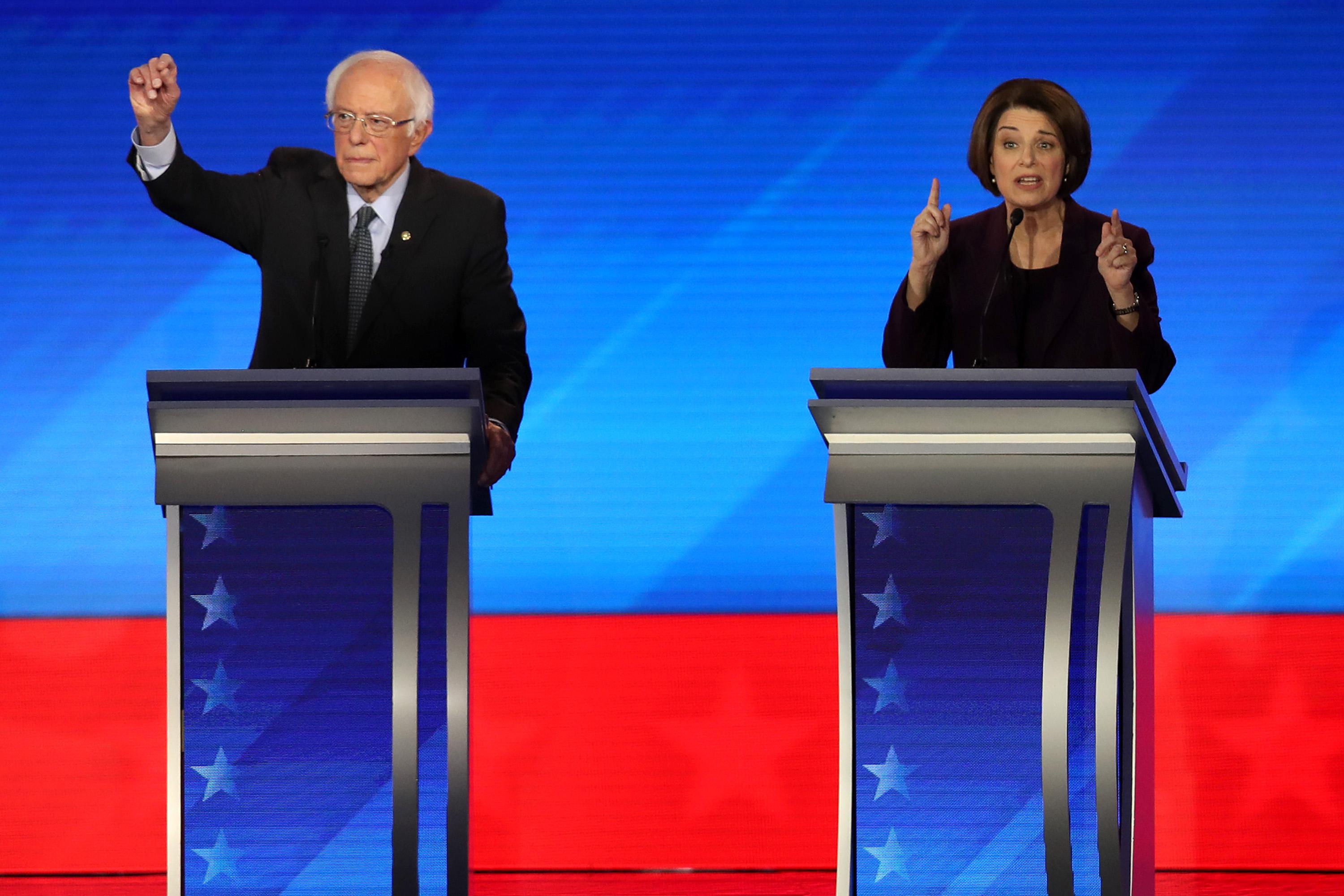 Bernie Sanders and Amy Klobuchar stand at their respective podiums while gesturing with their hands.