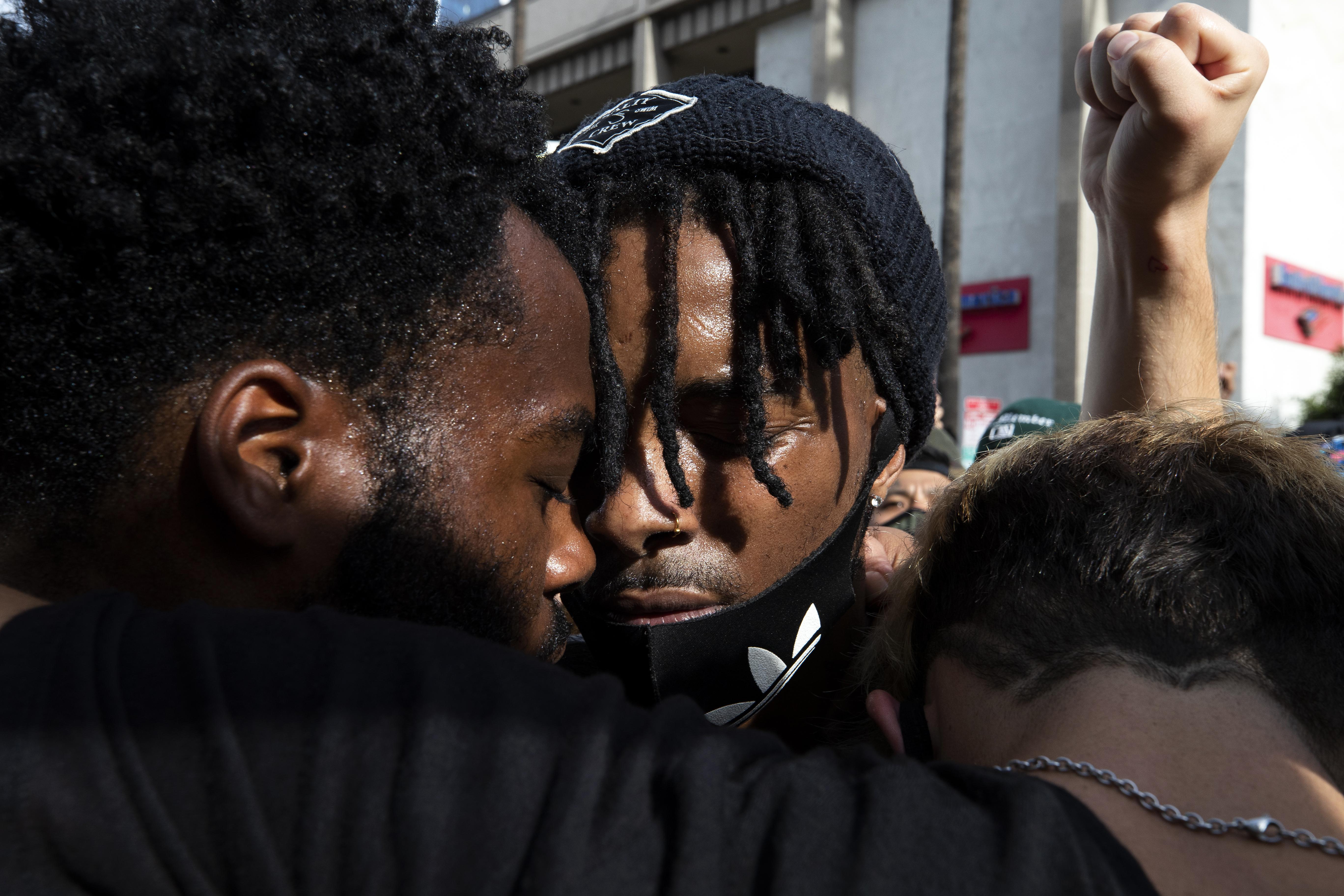 Two black protesters closing their eyes and putting their heads together as a white fist is held up in the shot.